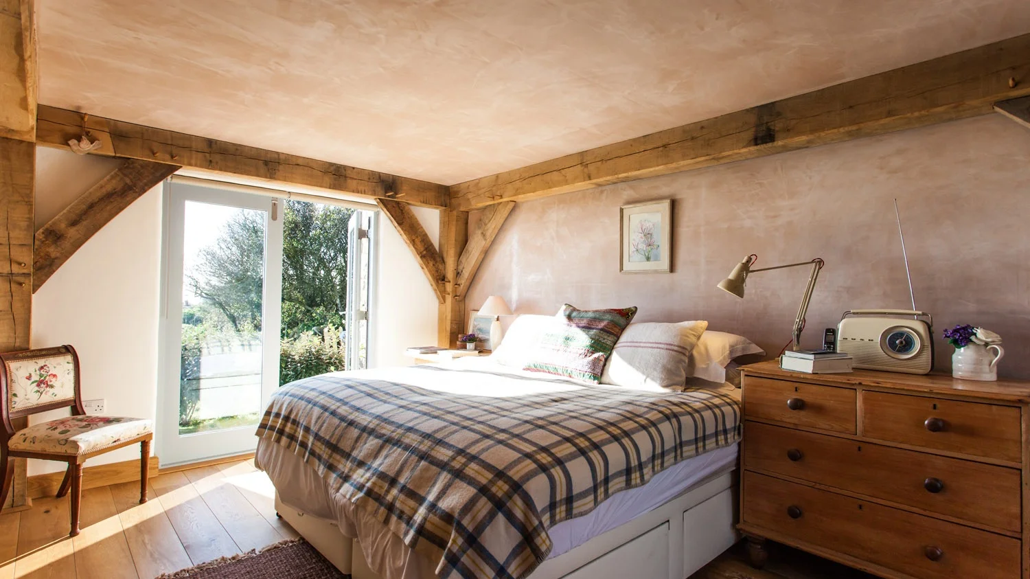 Cozy bedroom with a bed covered in plaid bedding, wooden dresser with books, radio, lamp, and flowerpot, large window showing outside greenery, floral chair in corner, and artwork on the wall.