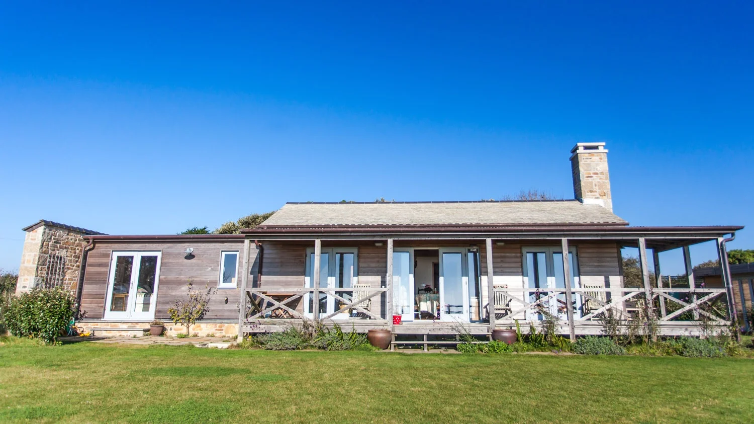 A house with a wooden exterior, multiple glass doors, a stone chimney, and a wrap-around porch, set against a clear blue sky.