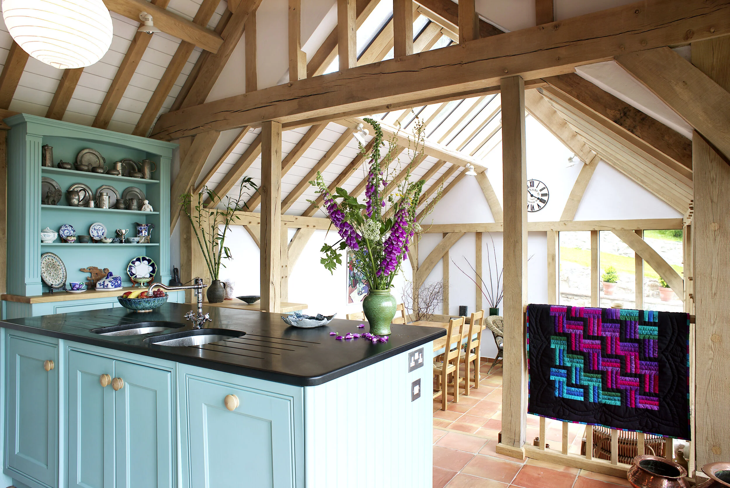 Inside a bright, open-concept kitchen with a rustic style, featuring exposed wooden beams, a turquoise cabinet filled with dishes, a black countertop with a large green floral vase, and a sitting/dining area with a wooden table and chairs, under skyl