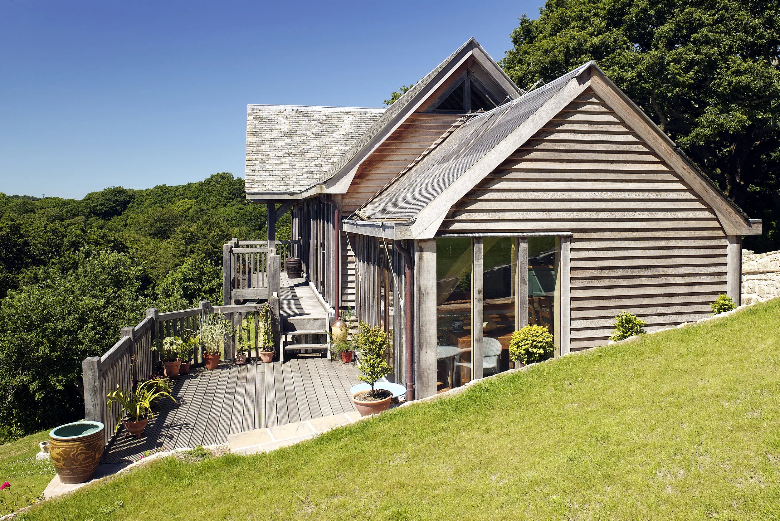 A rustic wooden house with a deck, surrounded by green trees and grass, with large windows and potted plants on the deck.