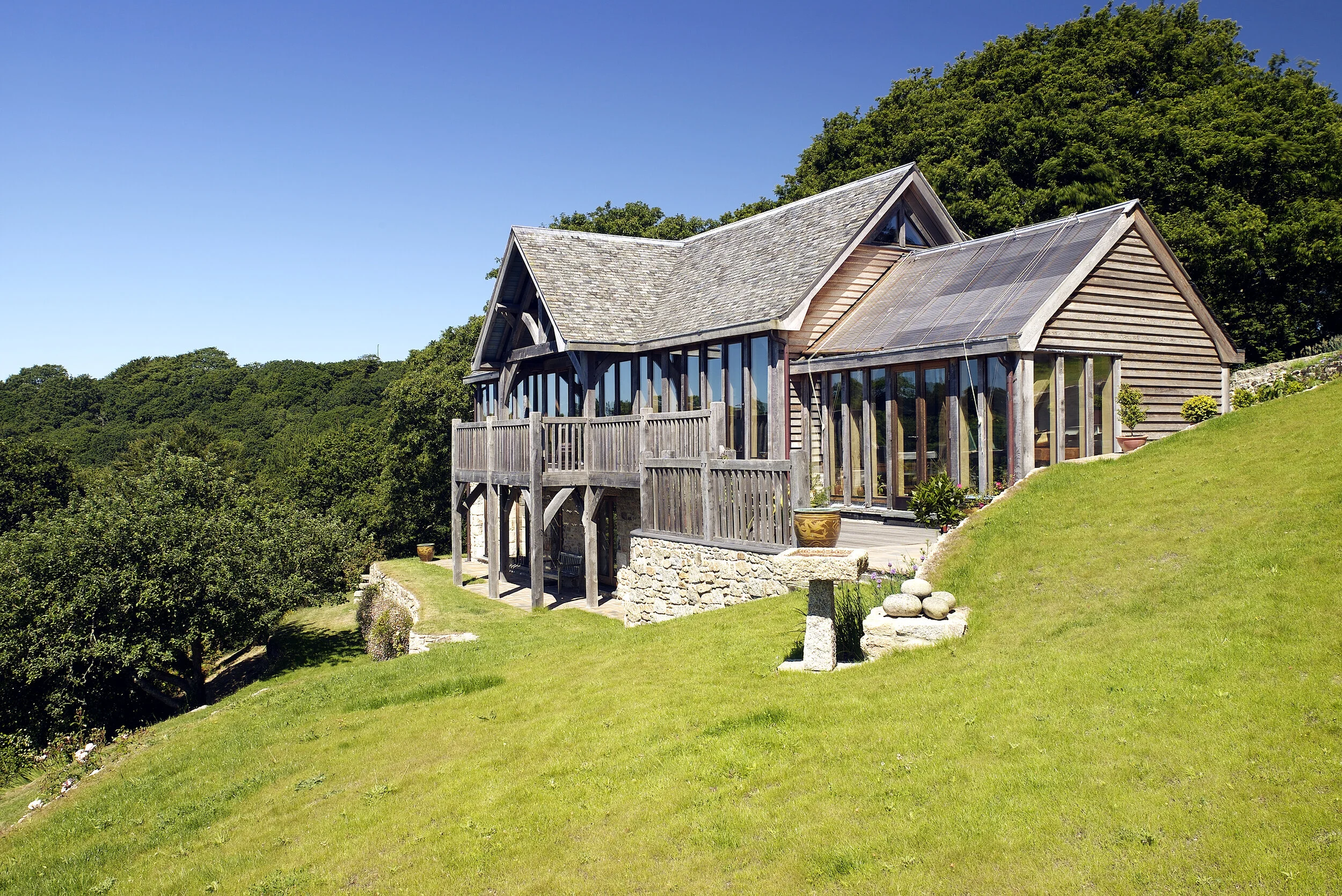 A house built on a hillside with a balcony on the second floor, large windows, and a sloped wooden roof, surrounded by green grass and trees, under a clear blue sky.