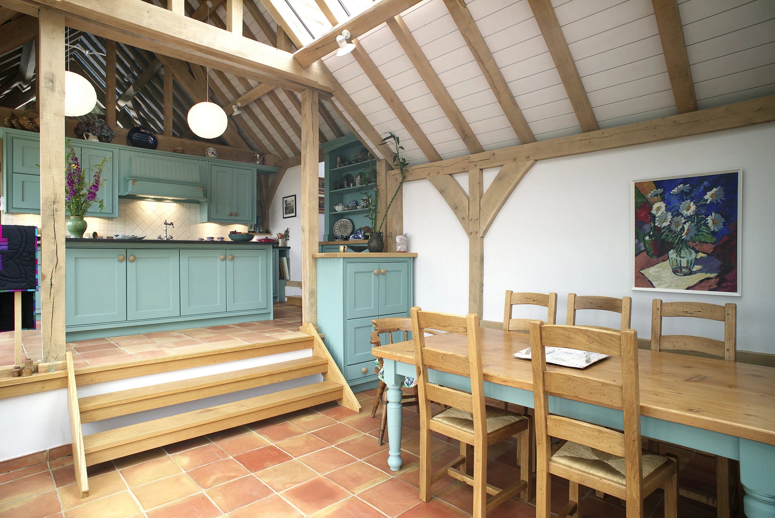 Interior of a kitchen and dining area with a wooden staircase, teal cabinets, a wooden dining table with six chairs, and artwork on the white walls.