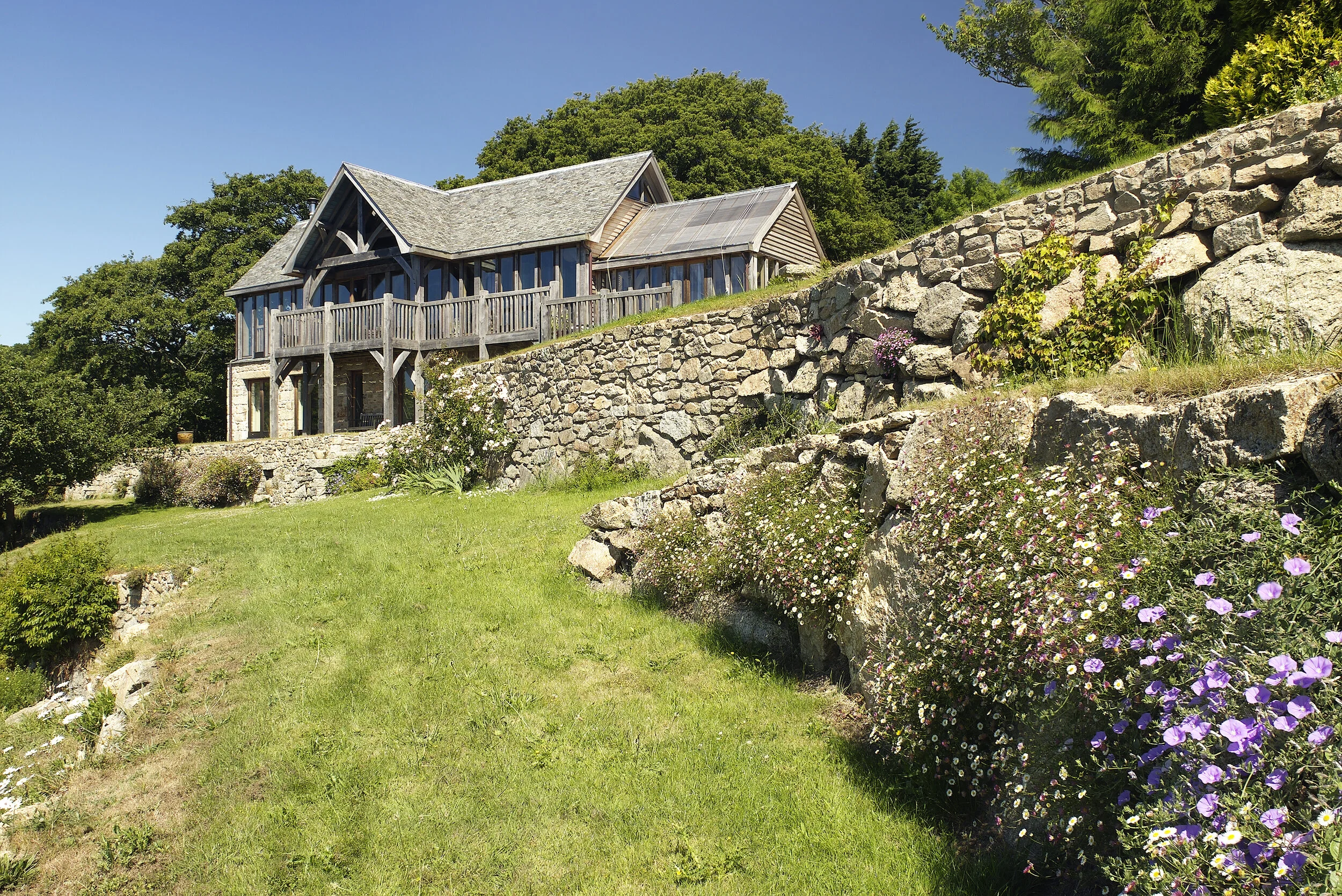 A two-story house with wooden siding and large windows, situated on a hillside with a stone retaining wall and surrounded by lush green trees and flowering plants.
