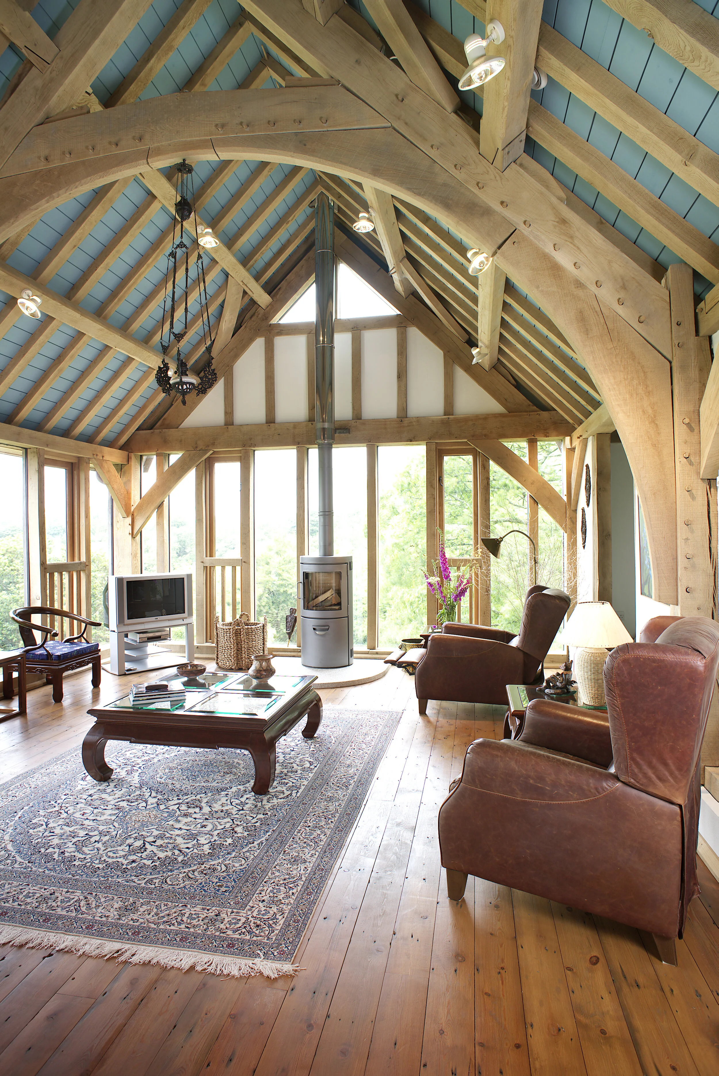 Interior view of a rustic living room with a high, vaulted ceiling, large windows, leather armchairs, wooden furniture, a patterned area rug, a wood stove, and natural wood beams.