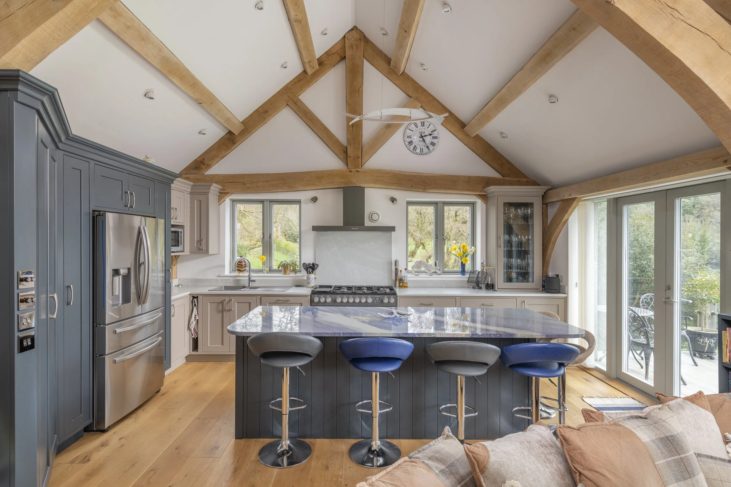Modern kitchen with exposed oak beams, large windows, a marble island, stainless steel refrigerator, and sliding glass doors leading to an outdoor patio.