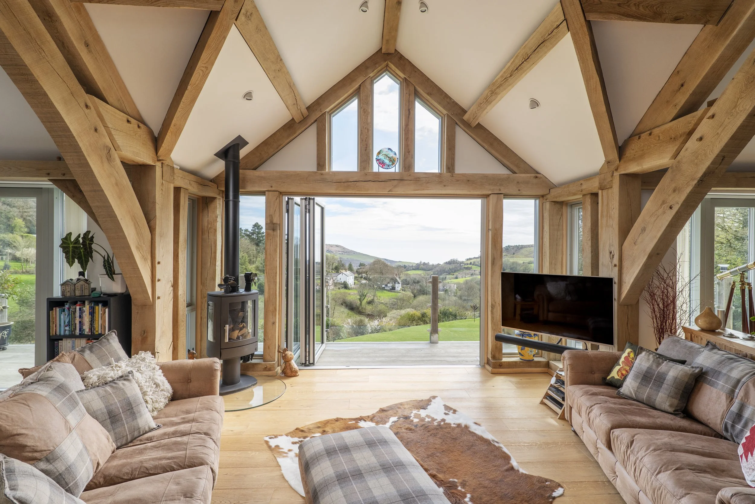Living room with oak beams, large windows, and a view of a green landscape. Contains two sofas with plaid and plain cushions, a cowhide rug, a TV, a wood stove, and decorations.