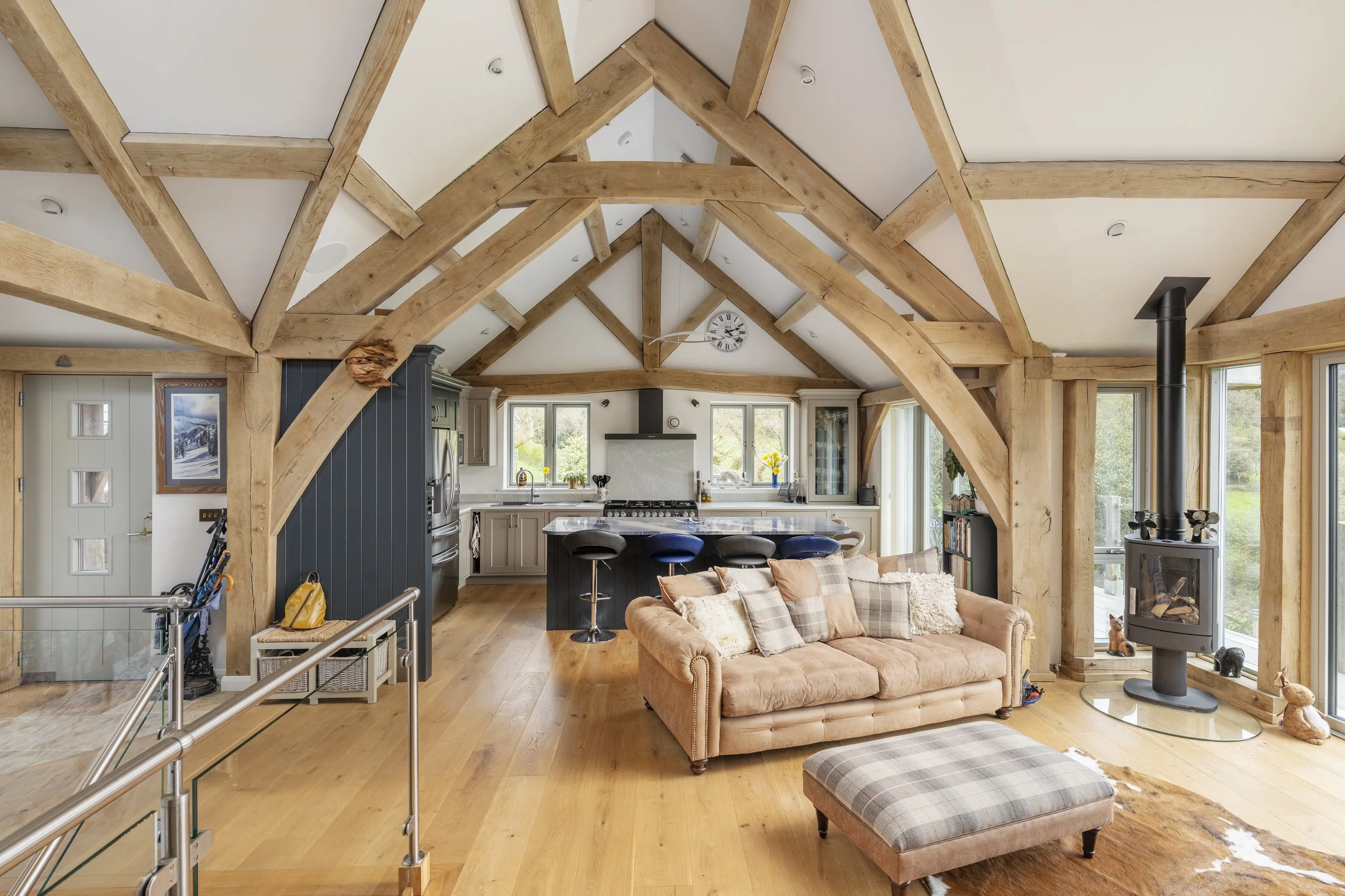 Open-concept living room and kitchen with exposed oak beams, tan couch with plaid pillows, and a modern wood stove near large windows.