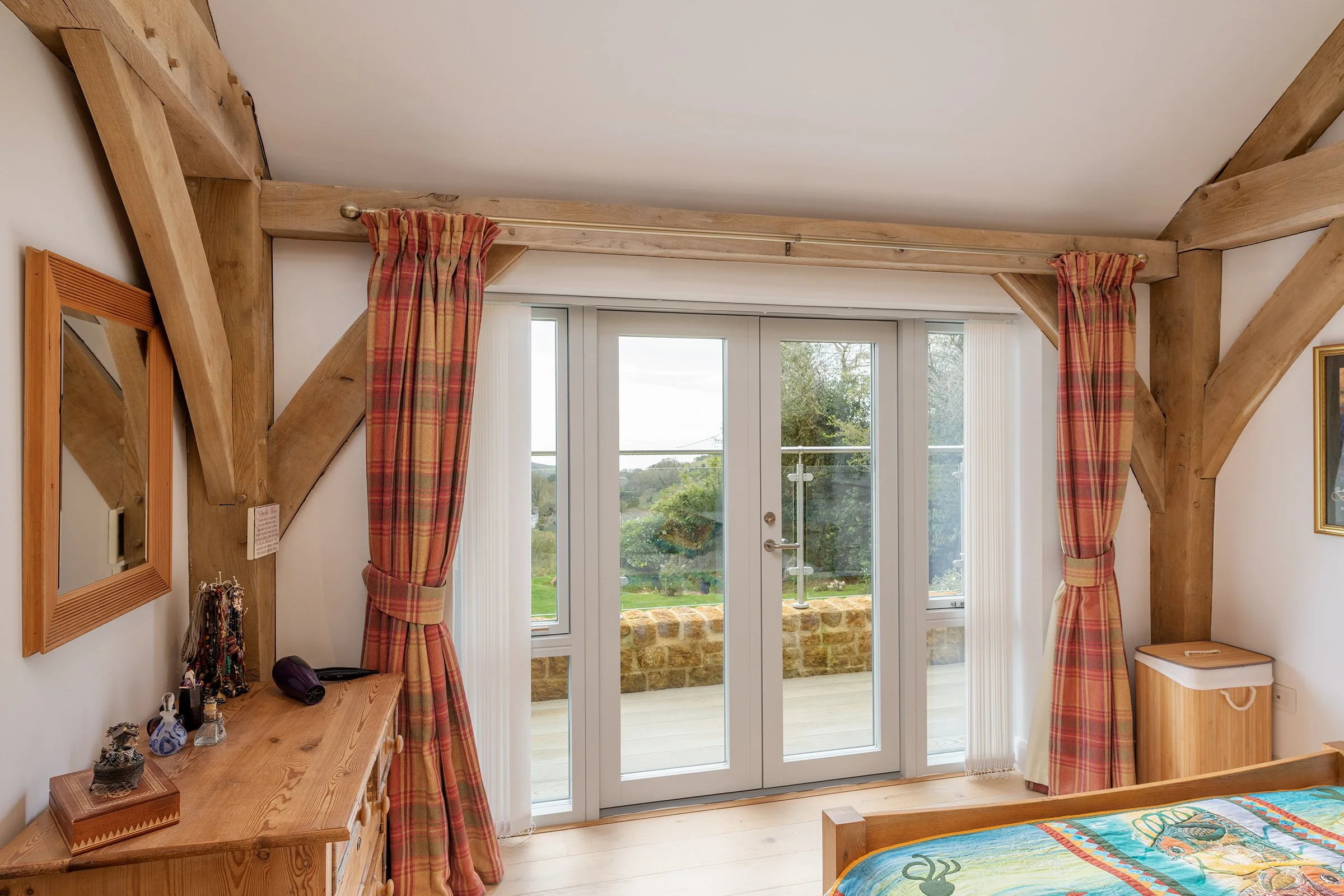 Room with oak beams, a glass door leading to an outdoor balcony, pink plaid curtains, and a bed with a colorful bedspread.