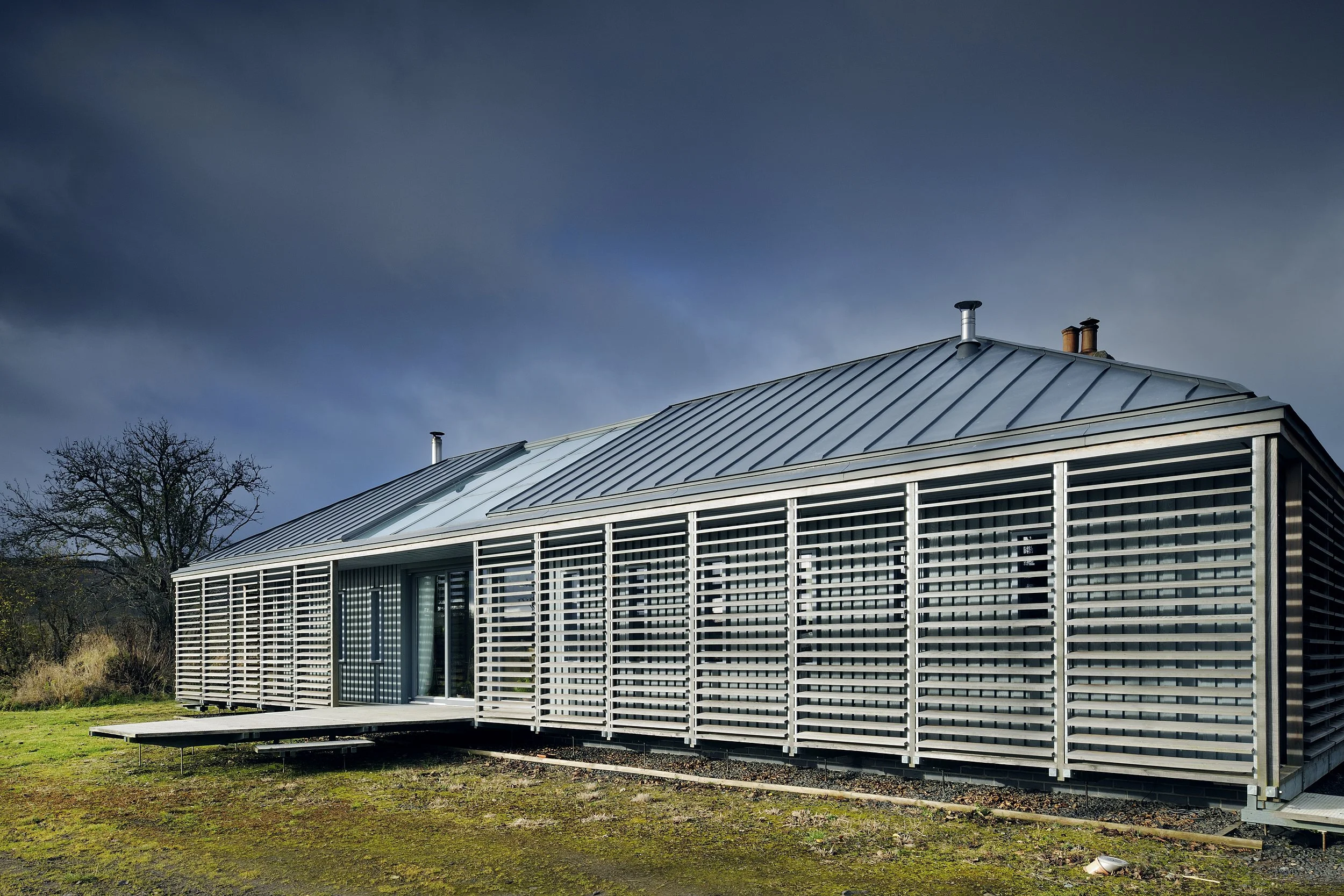 Modern house with metal siding and large louvered windows, situated on a grassy landscape under a cloudy sky.