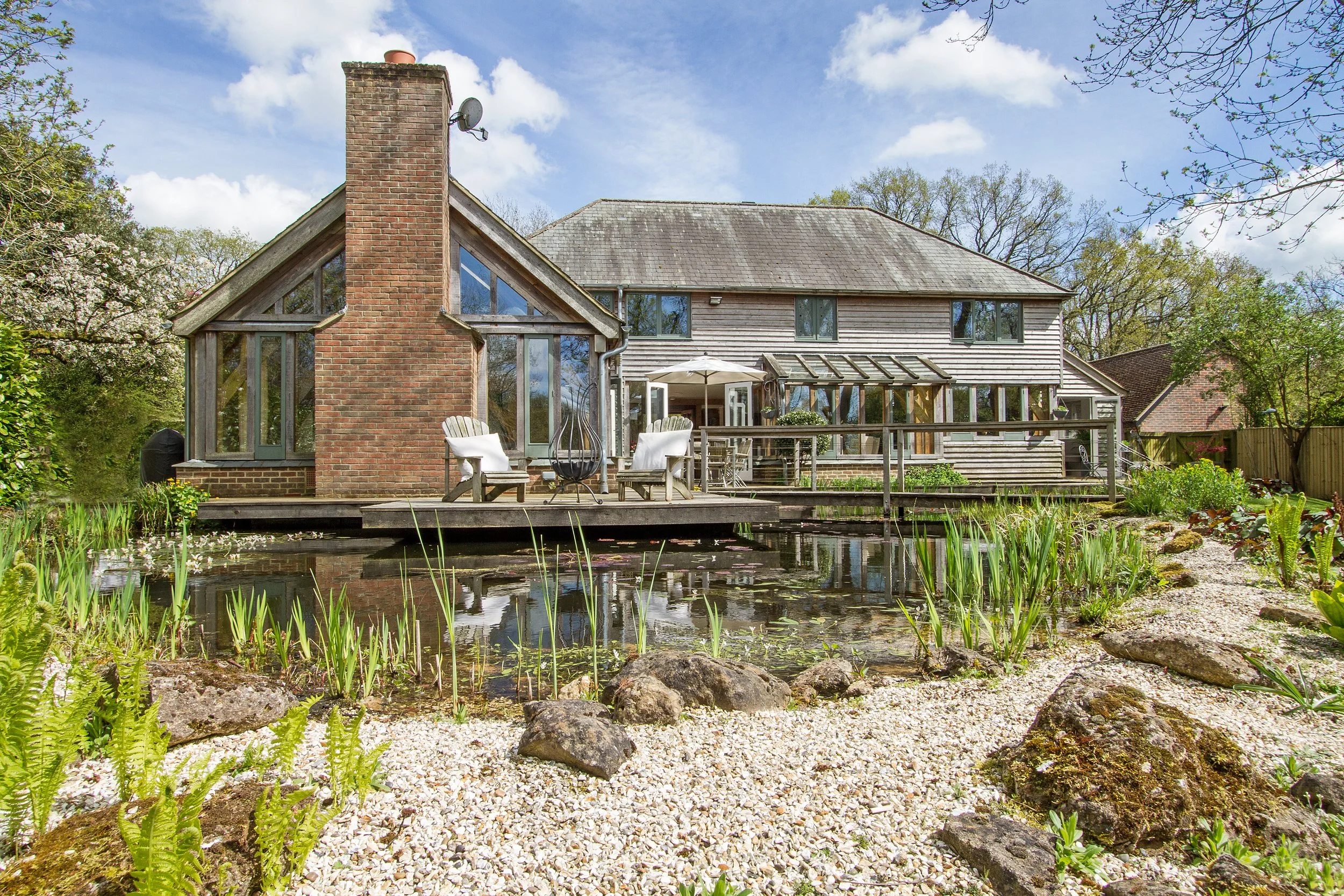 A backyard view of a two-story house with a wooden and brick exterior, large windows, and a chimney. There is a deck with outdoor furniture overlooking a pond with rocks and greenery.