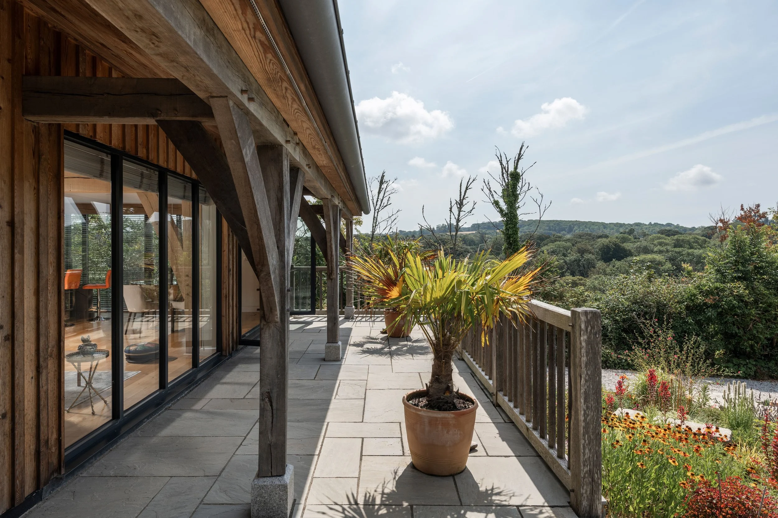 View of a balcony with large potted plant, wooden railing, and glass sliding doors leading inside a house. The sky is partly cloudy with scenic green hills in the distance.