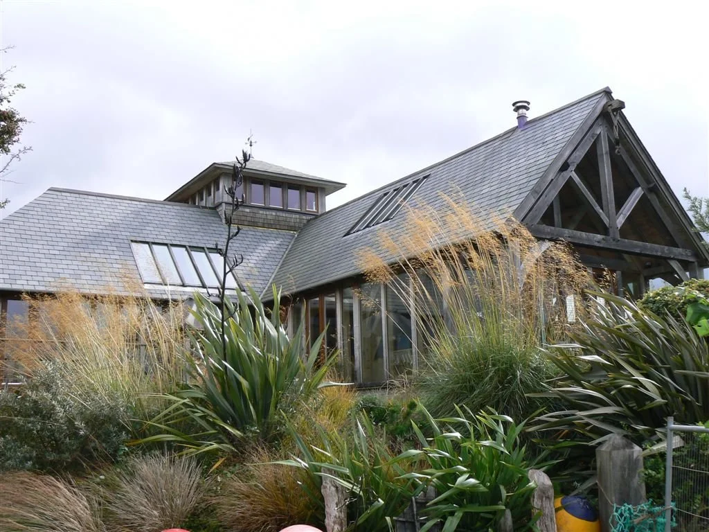 A modern house with gray slate roof, large skylights, and wooden beams, surrounded by lush garden with tall grasses and plants.