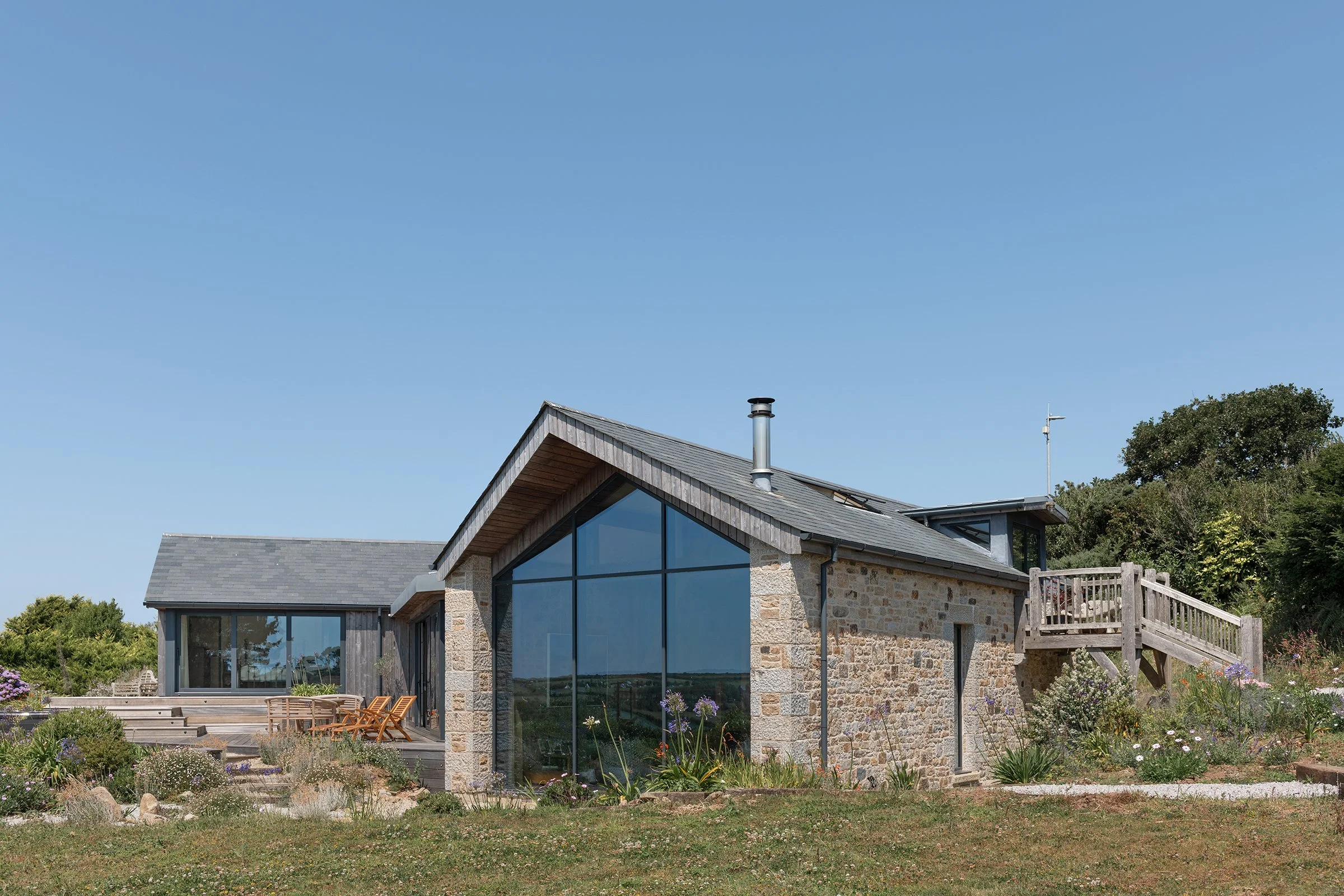 Modern house with large glass windows, stone and wood exterior, surrounded by a garden with flowers and shrubs under a clear blue sky.