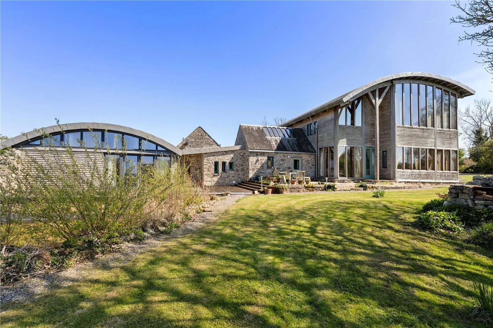 A modern house with large glass windows and wooden and stone exterior, set in a landscaped garden with green grass and plants, under a clear blue sky.