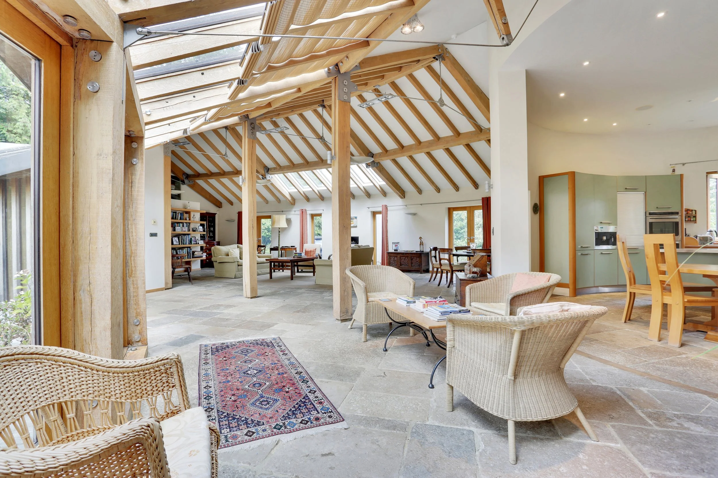 Spacious open-plan living room and kitchen with exposed timber beams, skylights, wicker furniture, stone flooring, and patterned area rug