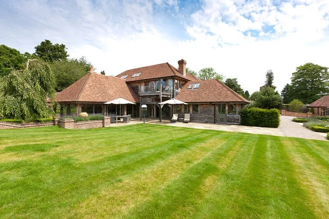 Large house with multiple roof sections and a spacious lawn in the foreground under a partly cloudy sky.