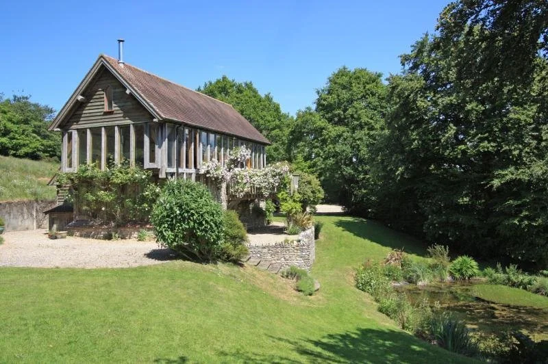 A wooden house with a sloped roof, surrounded by green trees and a garden, on a sunny day.