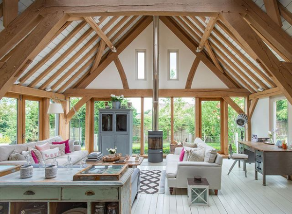 Interior of a spacious, bright living room with wooden beams, large windows, and modern furniture, including sofas, a coffee table, and a wood-burning stove.