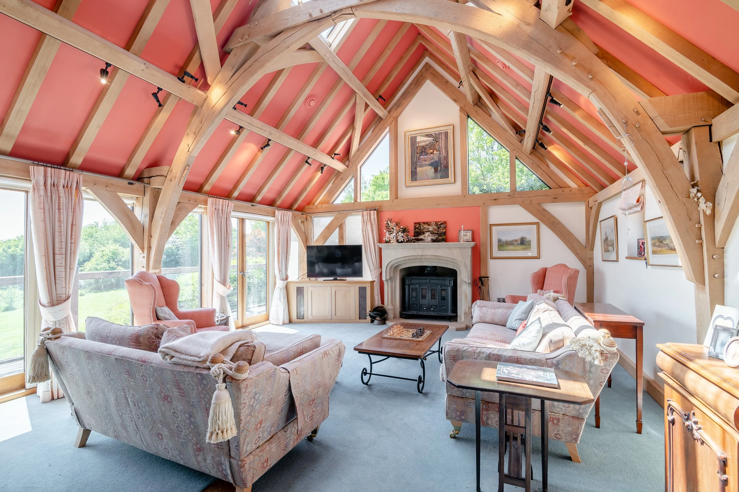 Living room with pink vaulted ceiling and exposed wooden beams, large windows with curtains, upholstered sofas, a fireplace, paintings on the walls, and a television.