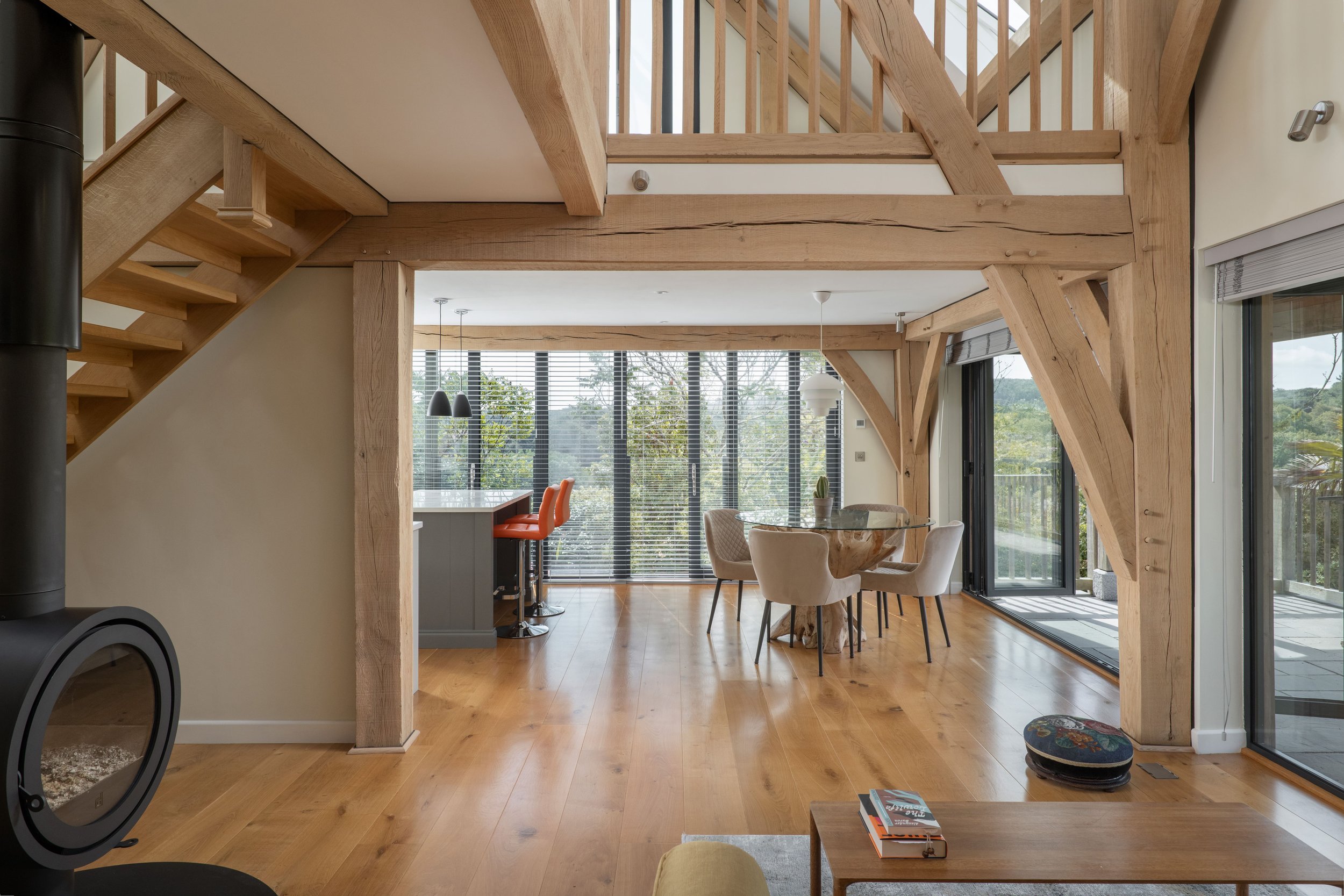 Open-concept interior of a home with wooden beams, large windows with blinds, a dining area with a round table and cream chairs, and a balcony with glass doors.