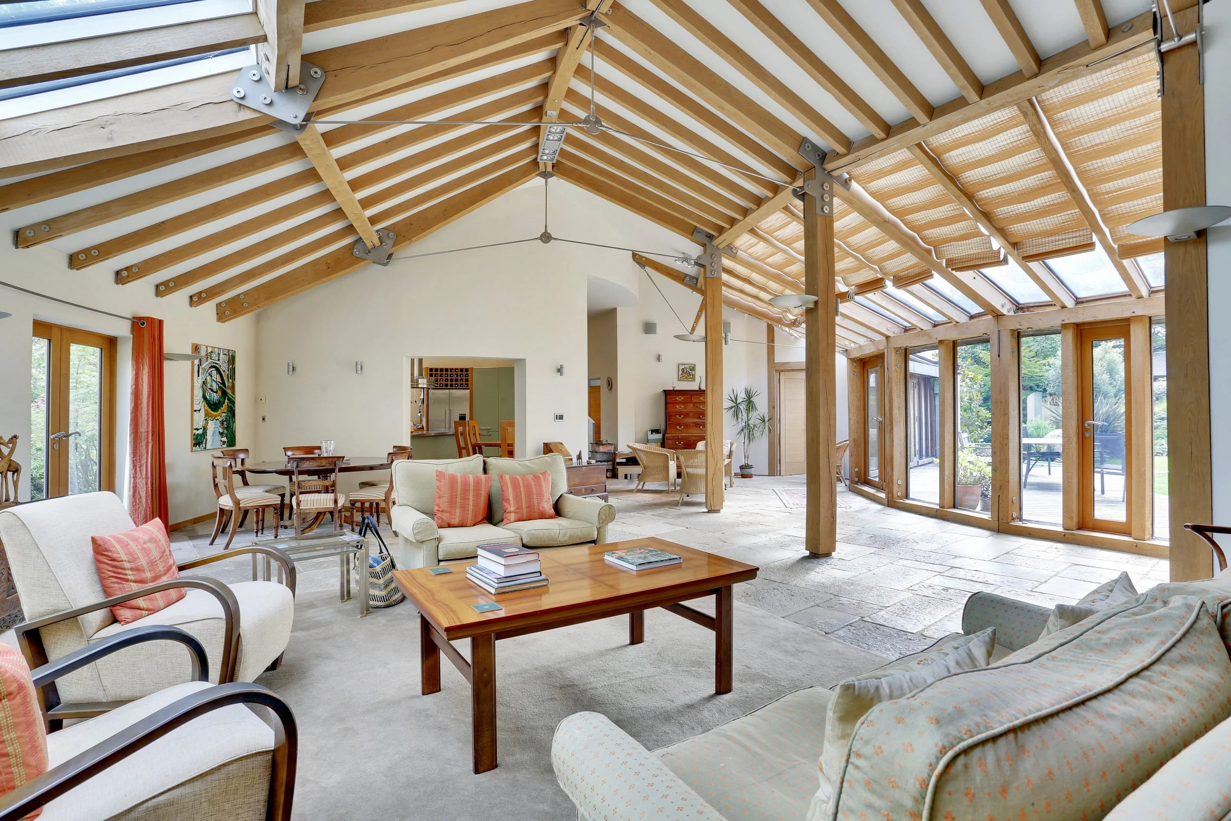 Bright open-plan living room with exposed timber rafters, skylights, glass doors, sofas, armchairs, dining table, and wooden coffee table