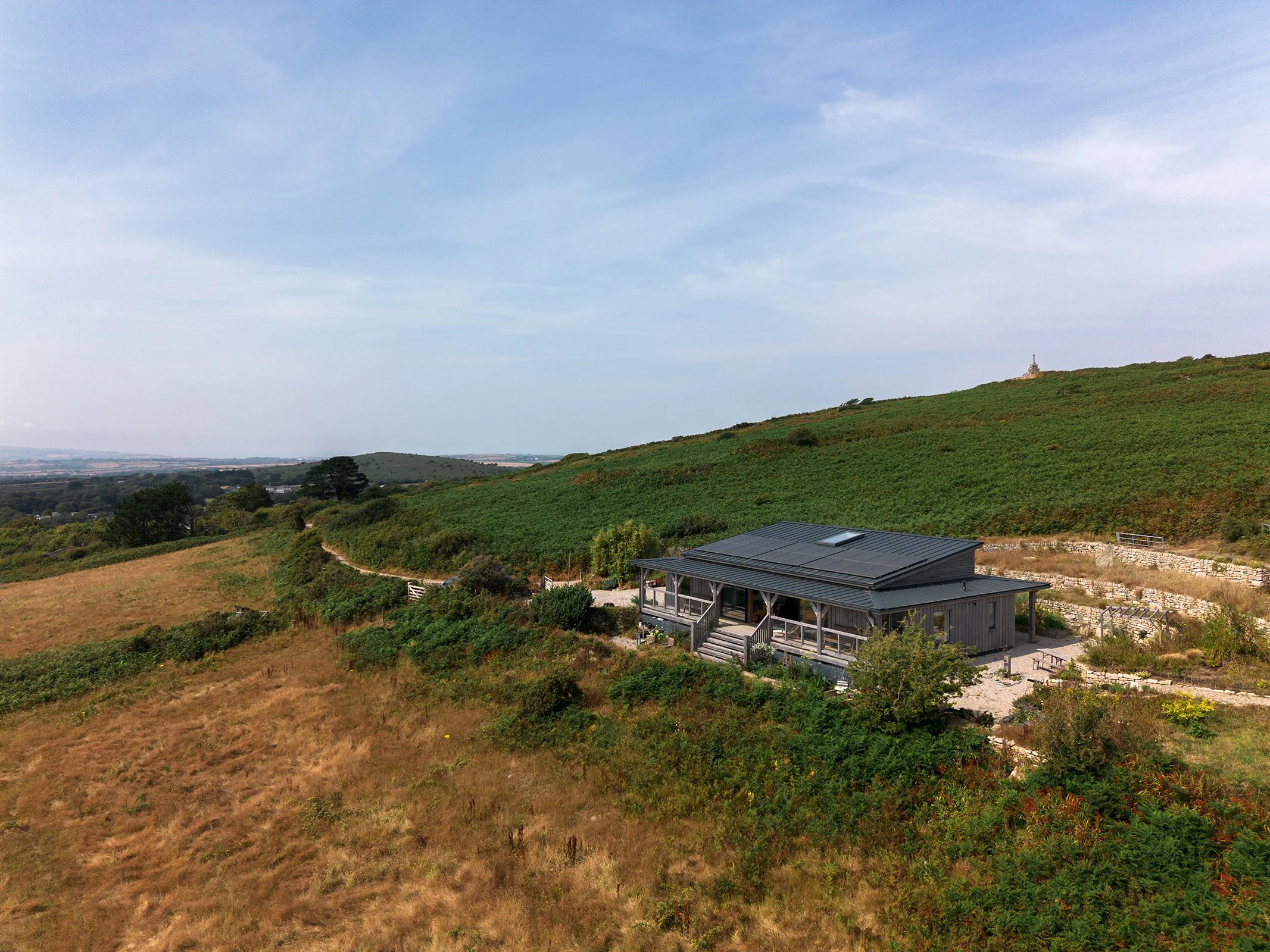 A landscape view of rolling green hills with a small modern house in the foreground, a church steeple on the horizon, under a partly cloudy sky.