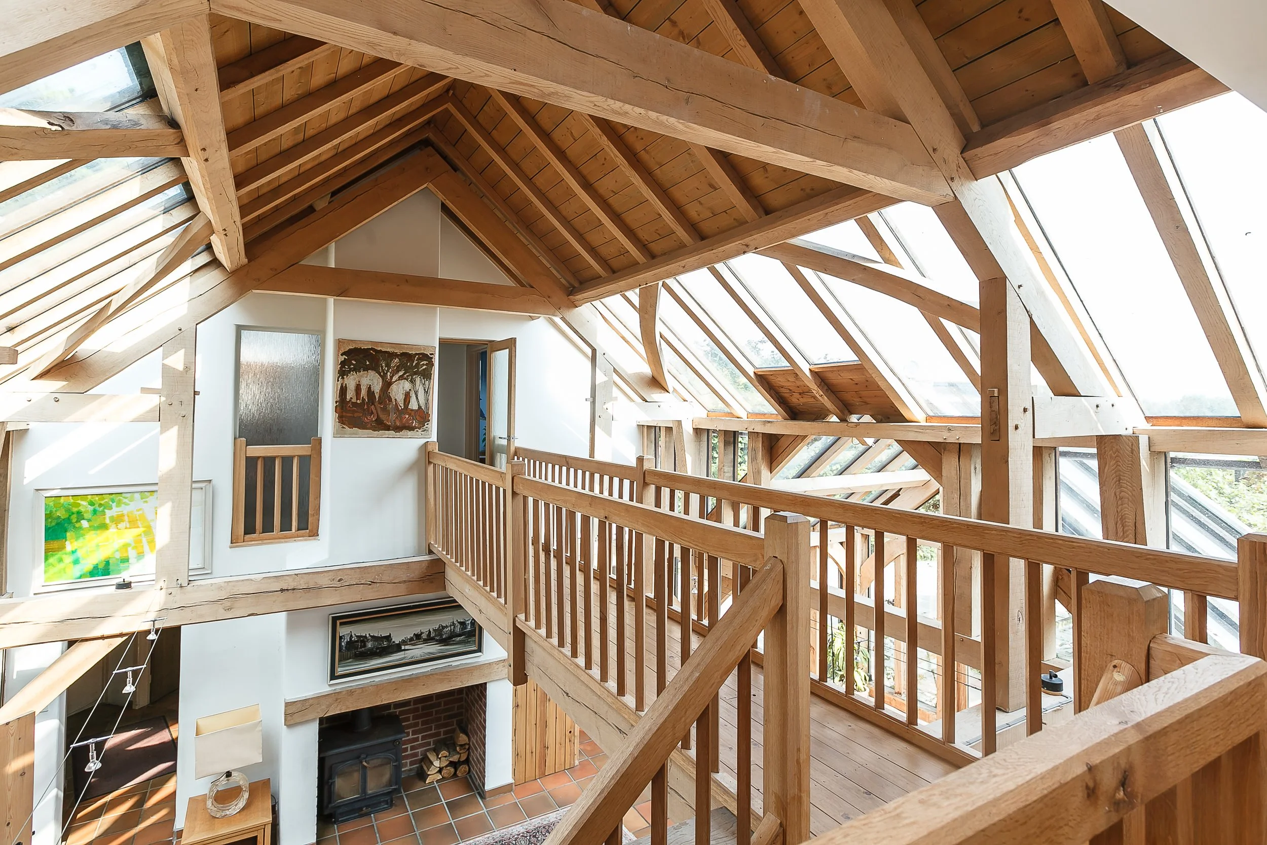 Interior view of a house with wooden beams and large skylight windows in the sloped ceiling, overlooking a living space with a fireplace, artwork on the wall, and a balcony railing.