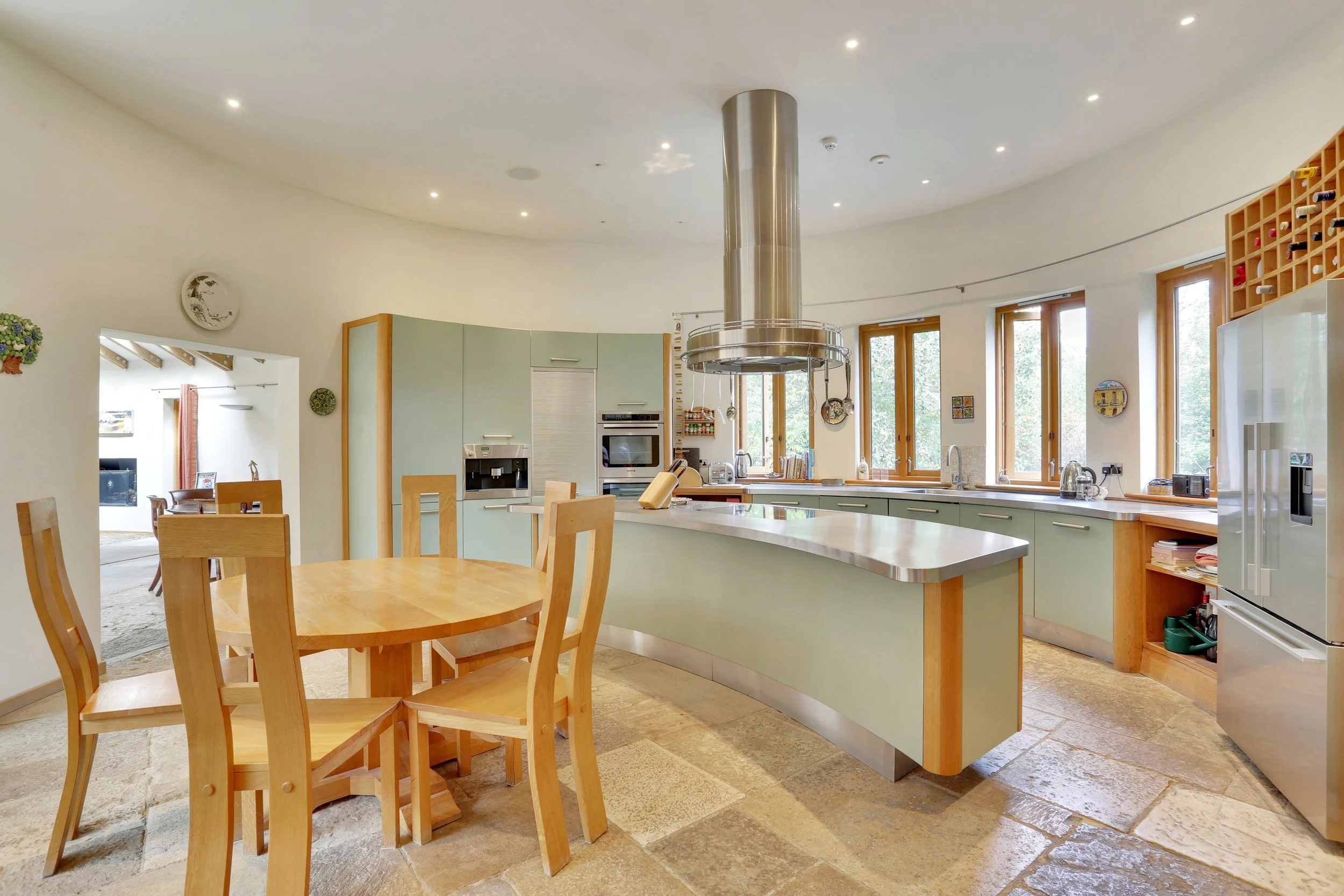 Spacious modern kitchen with a central island, stainless steel range hood, wooden dining table, large windows, and green cabinetry