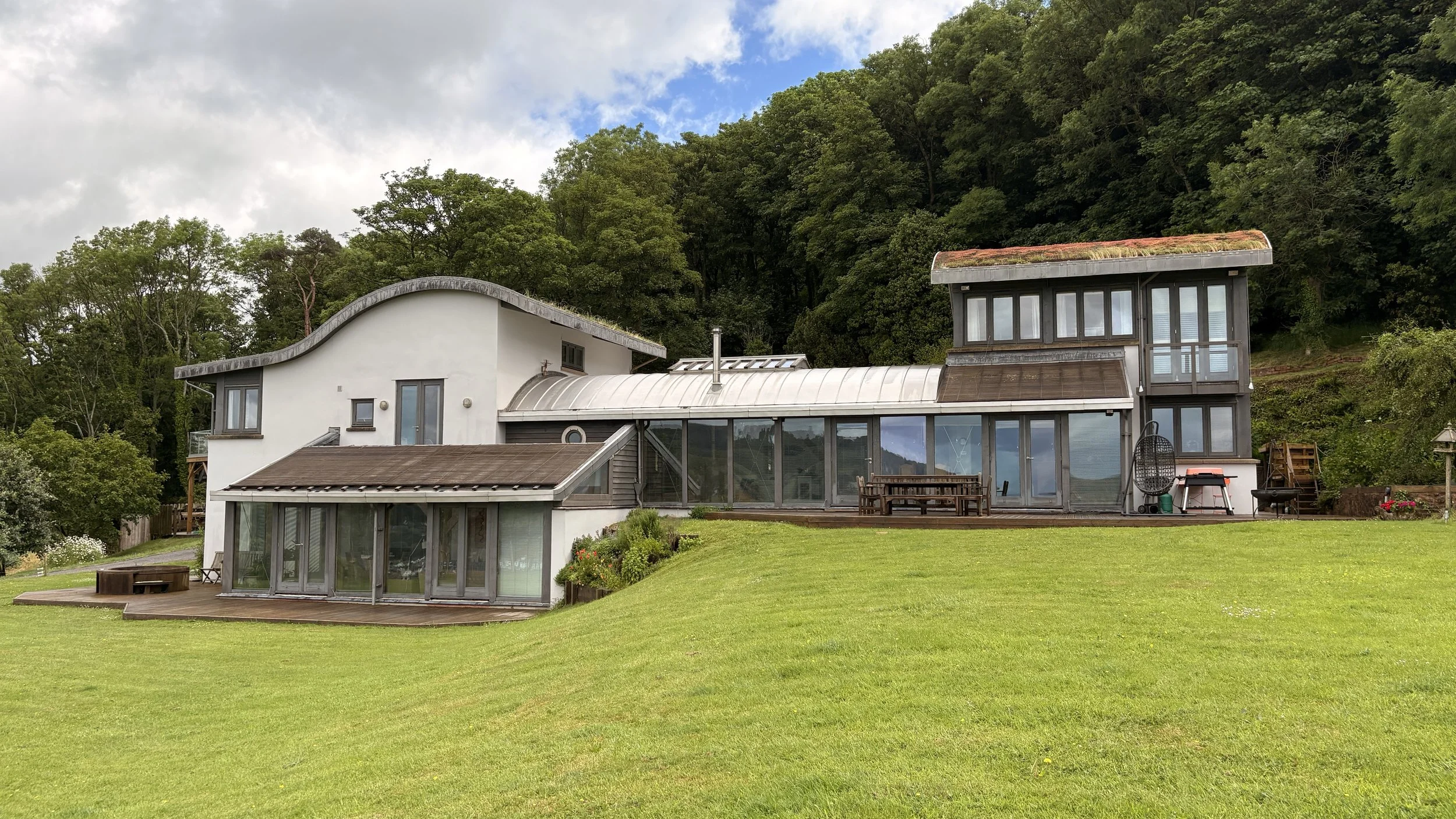 Modern house with multiple sections and large windows, situated on a grassy lawn with a wooded hillside in the background.
