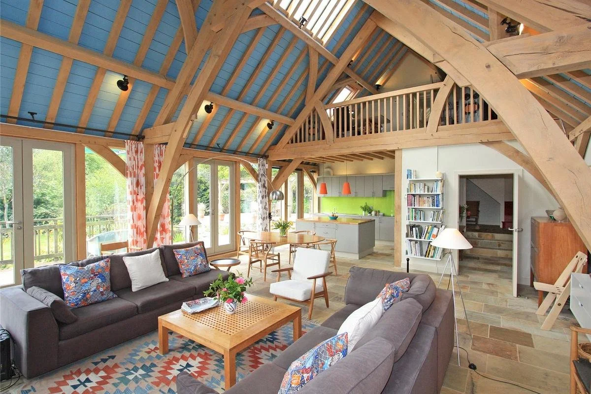 Inside view of a modern, open-concept living room and kitchen with oak beams, blue ceiling, large windows, and colorful decor.