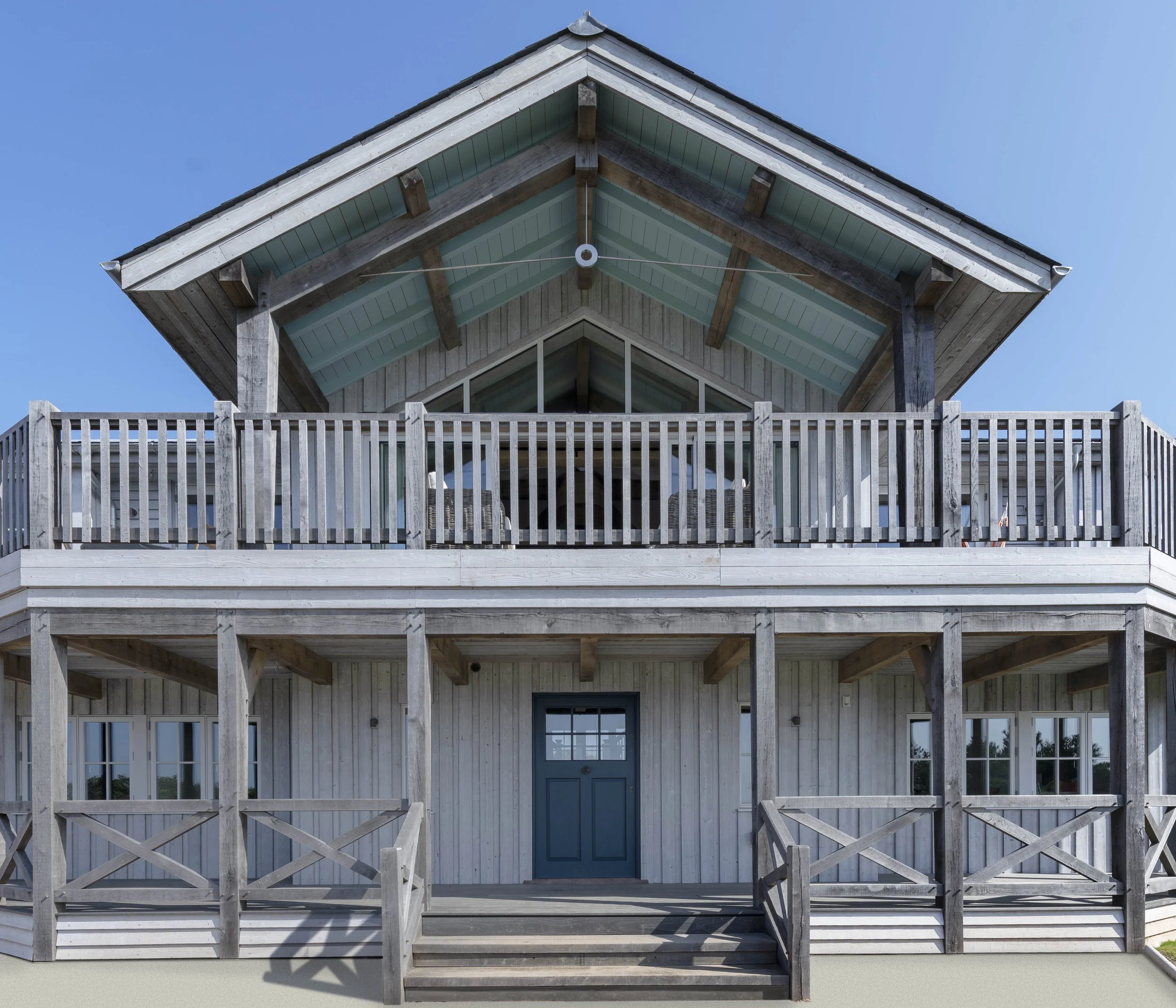 A two-story timber framedhouse with balconies on the upper levels, a central front door, and a blue door. The house has a large overhanging roof and is set against a clear blue sky.