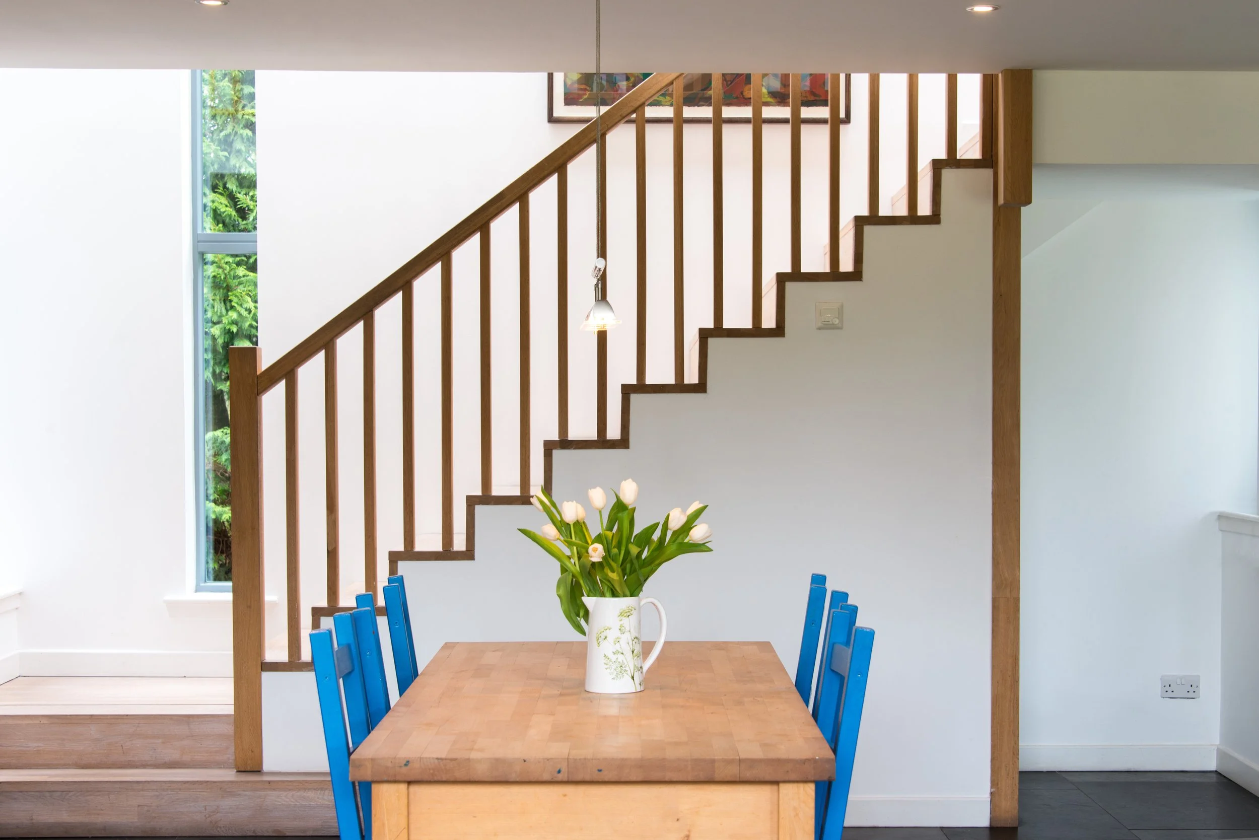 A dining area with a wooden table, blue chairs, and a white vase with white tulips. A staircase with wooden railings is in the background, along with a window and framed artwork.