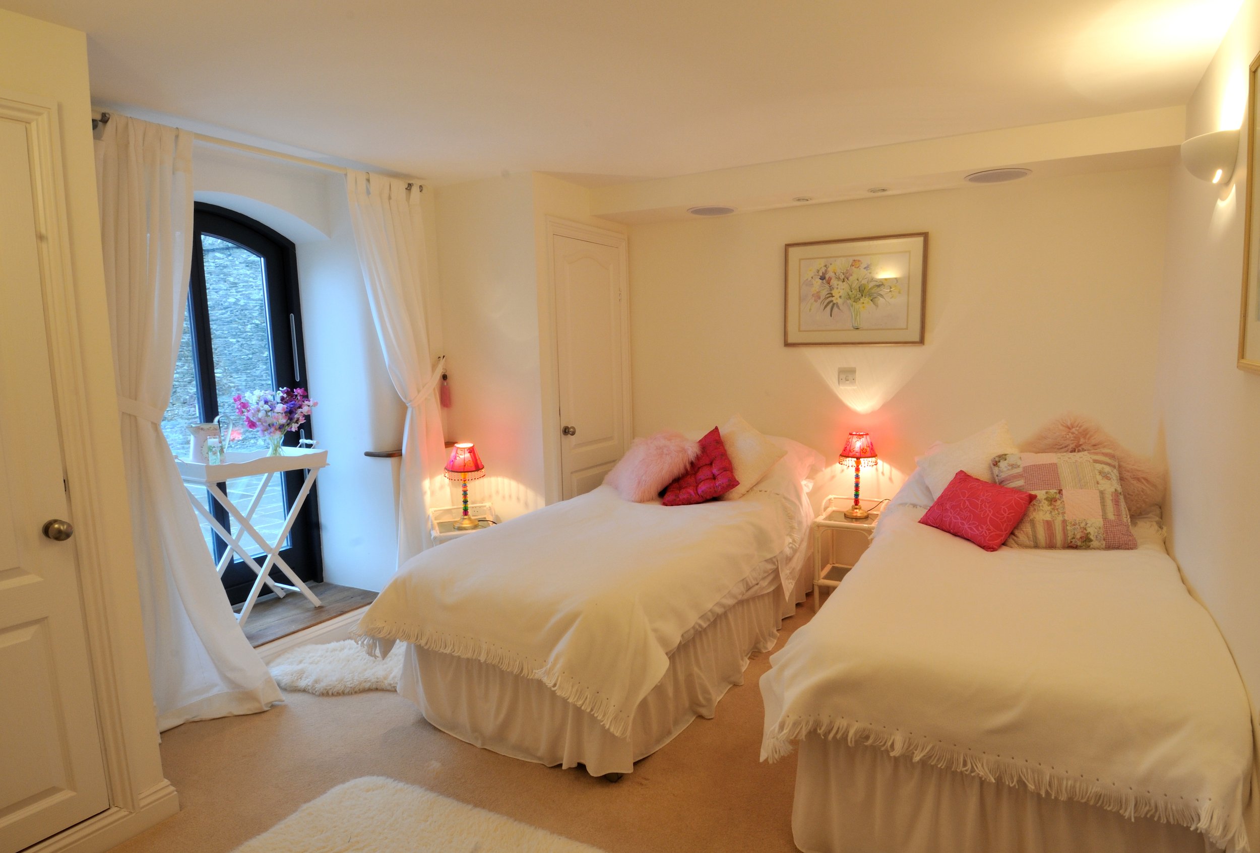 A cozy bedroom with two beds, decorated with pink and patterned pillows. There are nightstands with small red lamps, a framed floral picture on the wall, a window with white curtains, and a small balcony with flowers outside.