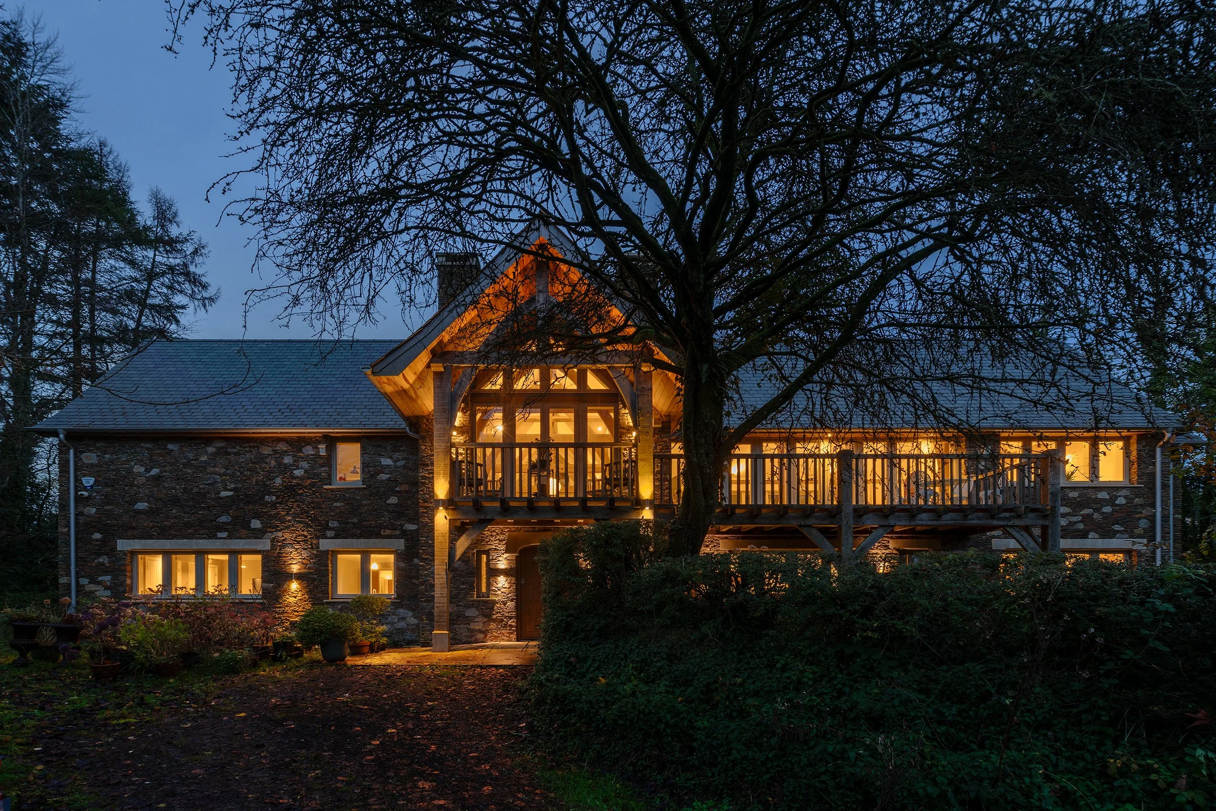 A large two storey stone house at duskwith the lights on and a tree, with no leaves, in front