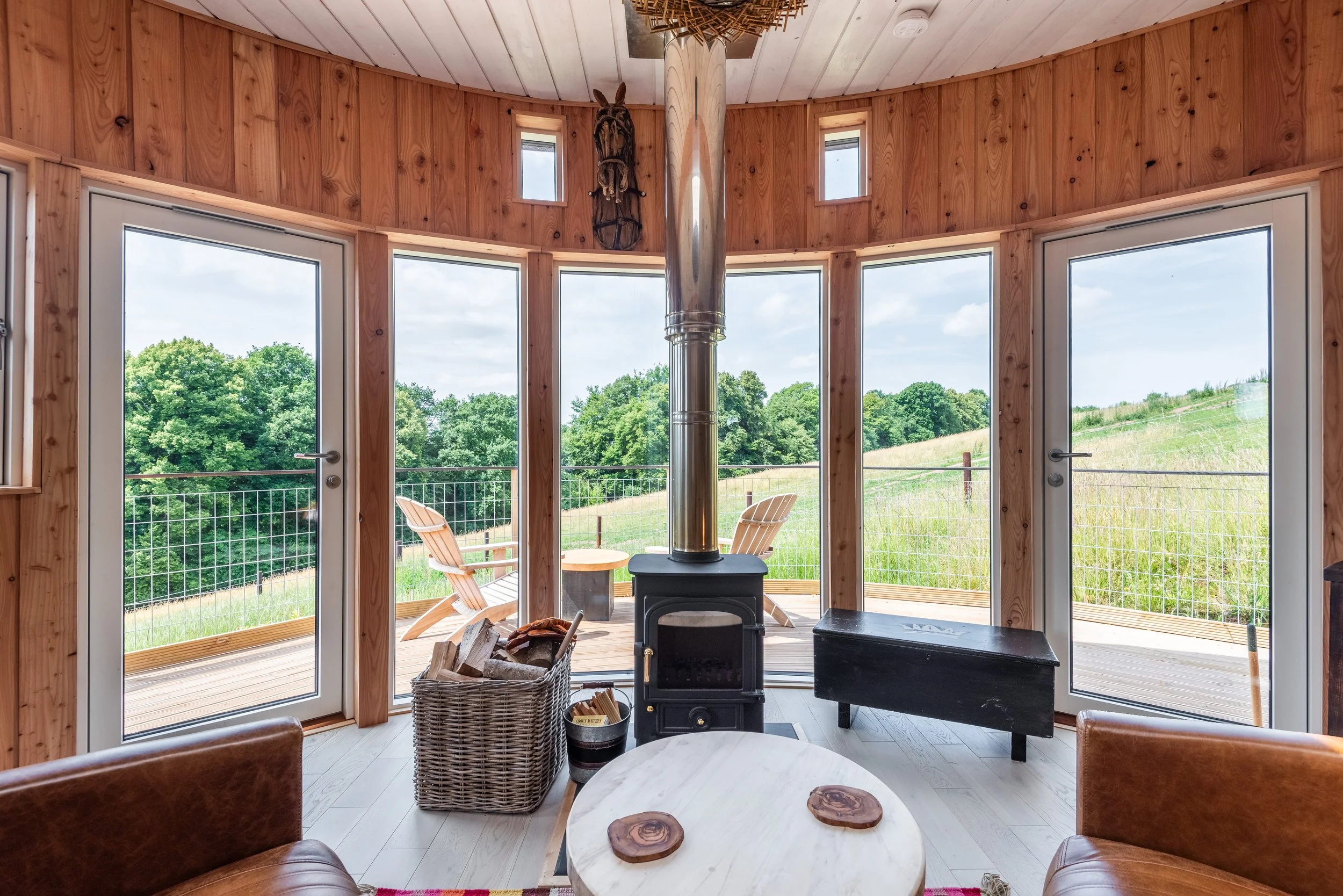 Interior of a cozy wooden cabin with a wood stove, circular white table, leather chairs, and large windows showing a green outdoor landscape with trees and rolling hills.