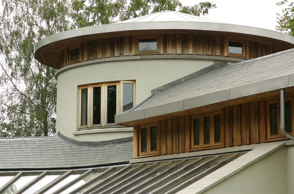Modern round-roofed building with wood-clad upper walls, large windows, and gray slate roofs surrounded by trees