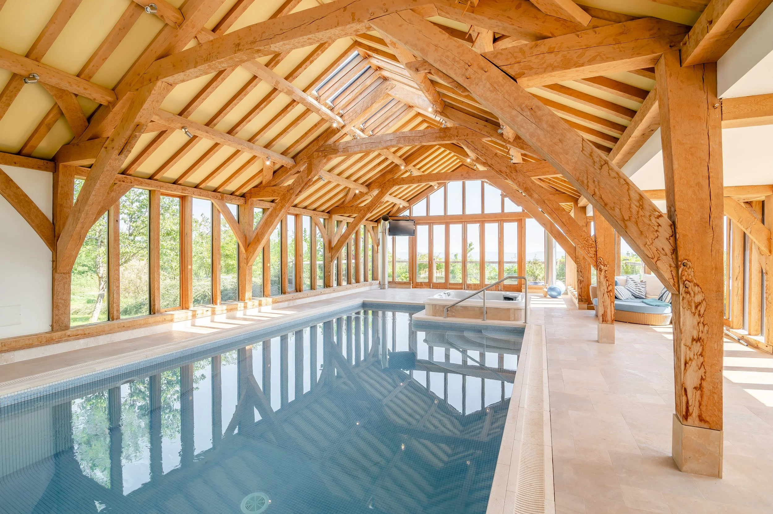 Indoor swimming pool area with wooden beams and large windows overlooking greenery, with a hot tub and seating area.