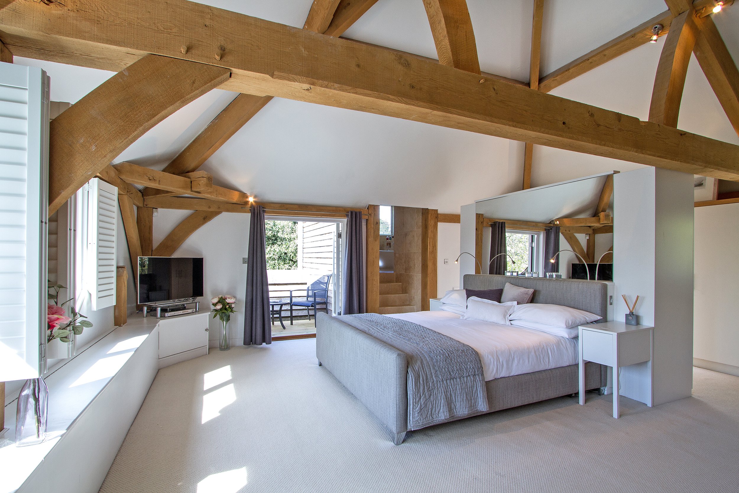 Modern bedroom with a sloped ceiling, large bed with white bedding, a TV on a white stand, a glass vase with flowers, and a balcony with outdoor seating visible through windows.
