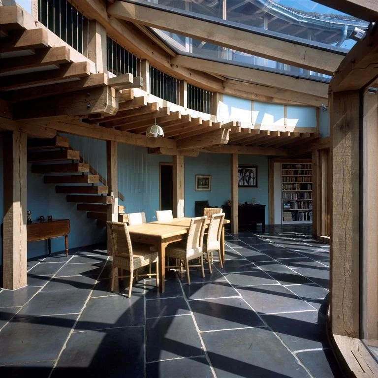 Open indoor space with black tiled floor, wooden staircase, and a wooden dining table with six chairs. There's a light fixture hanging above the table, a bookshelf, and some framed pictures on the blue walls.