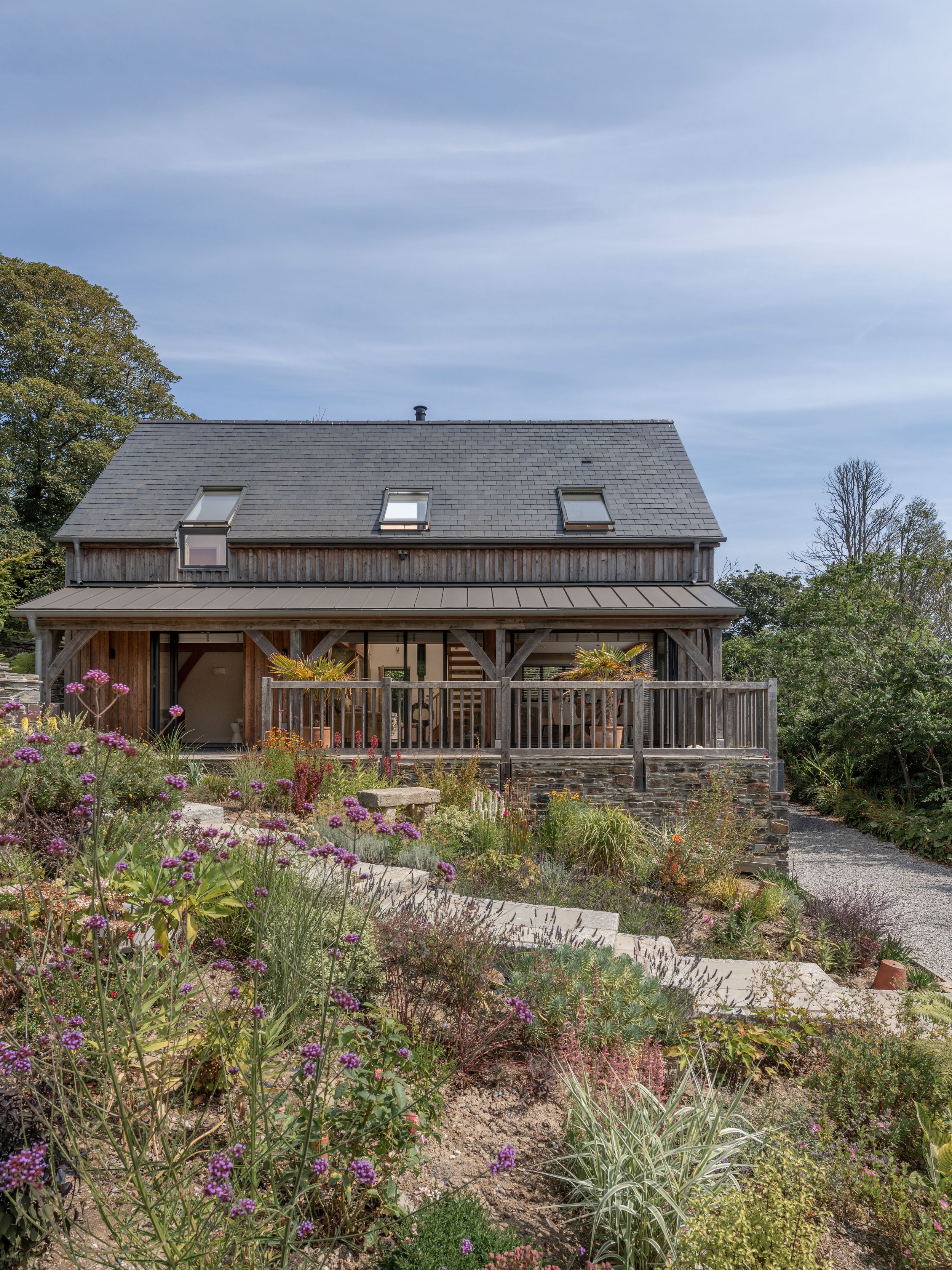 A house with a gray roof and skylights, surrounded by a colorful garden with various flowers and plants, and trees in the background under a cloudy sky.