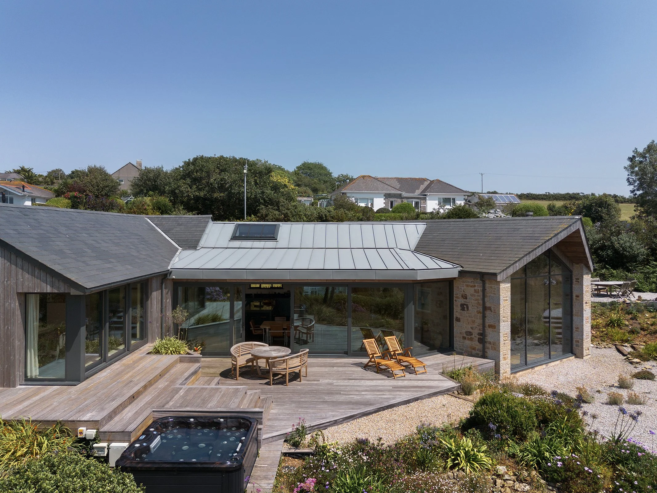 A modern house with large glass windows, a wooden deck with outdoor furniture, surrounded by gardens and rooftops, under a clear blue sky.