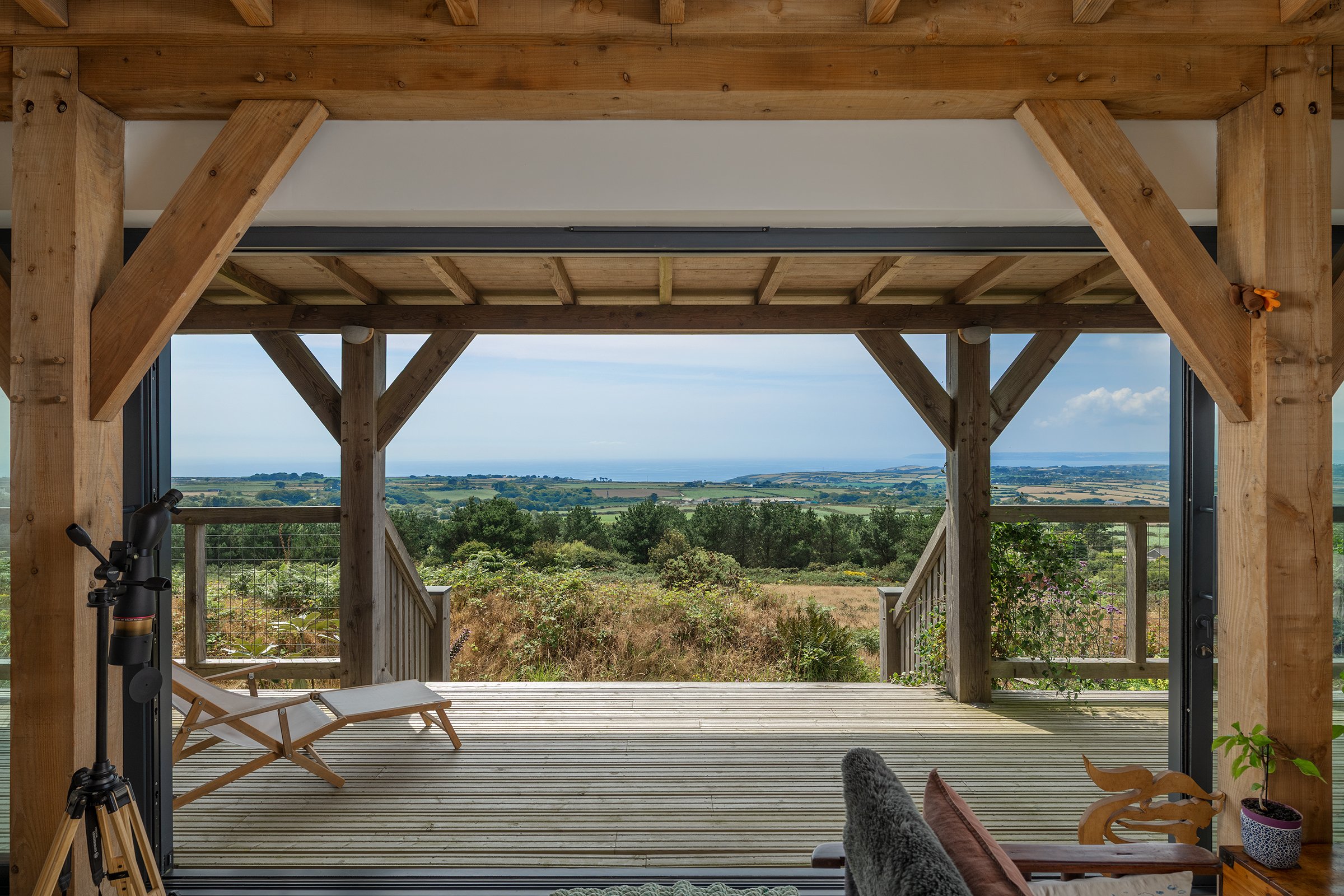 Wooden house with an open wall view of a landscape with green fields, trees, and a body of water in the distance under a partly cloudy sky.