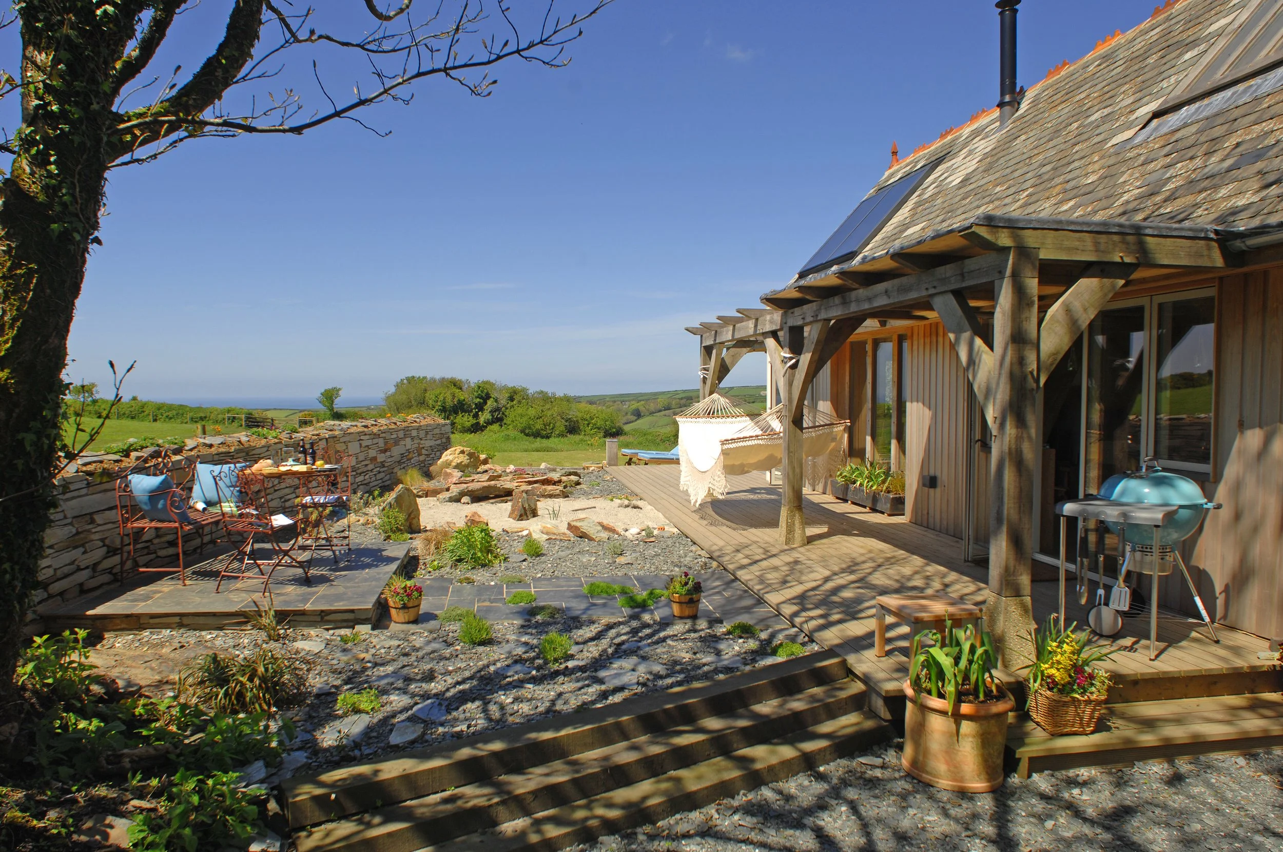 A cozy outdoor patio area with a stone seating wall, couple of chairs, and a small table set for outdoor dining on a bright sunny day. A wooden deck extends from a rustic house with solar panels on the roof. A hammock hangs between two posts on the d