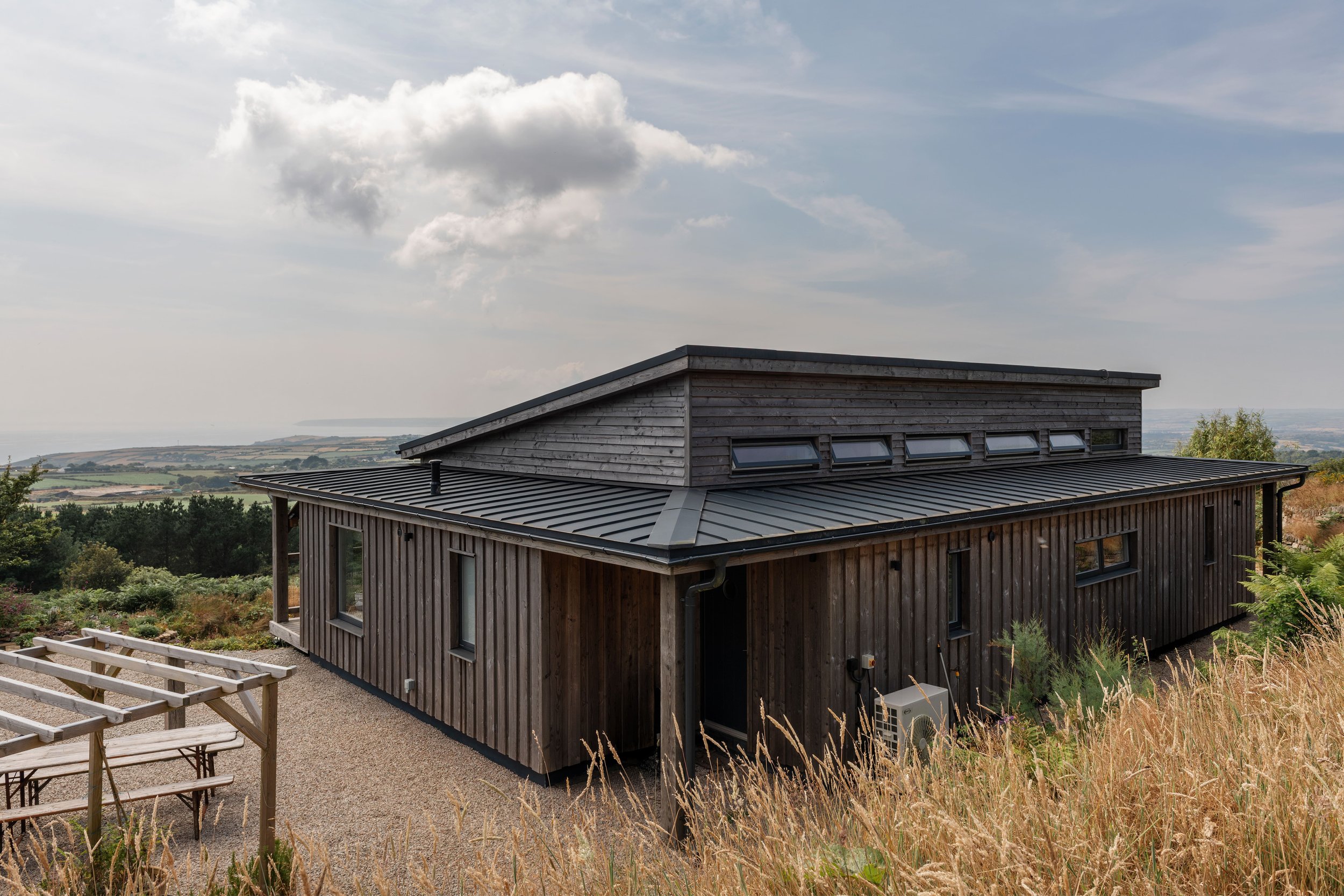 A modern wooden house with a sloped metal roof and multiple skylights, situated on a hillside overlooking a landscape with fields and trees under a partly cloudy sky.