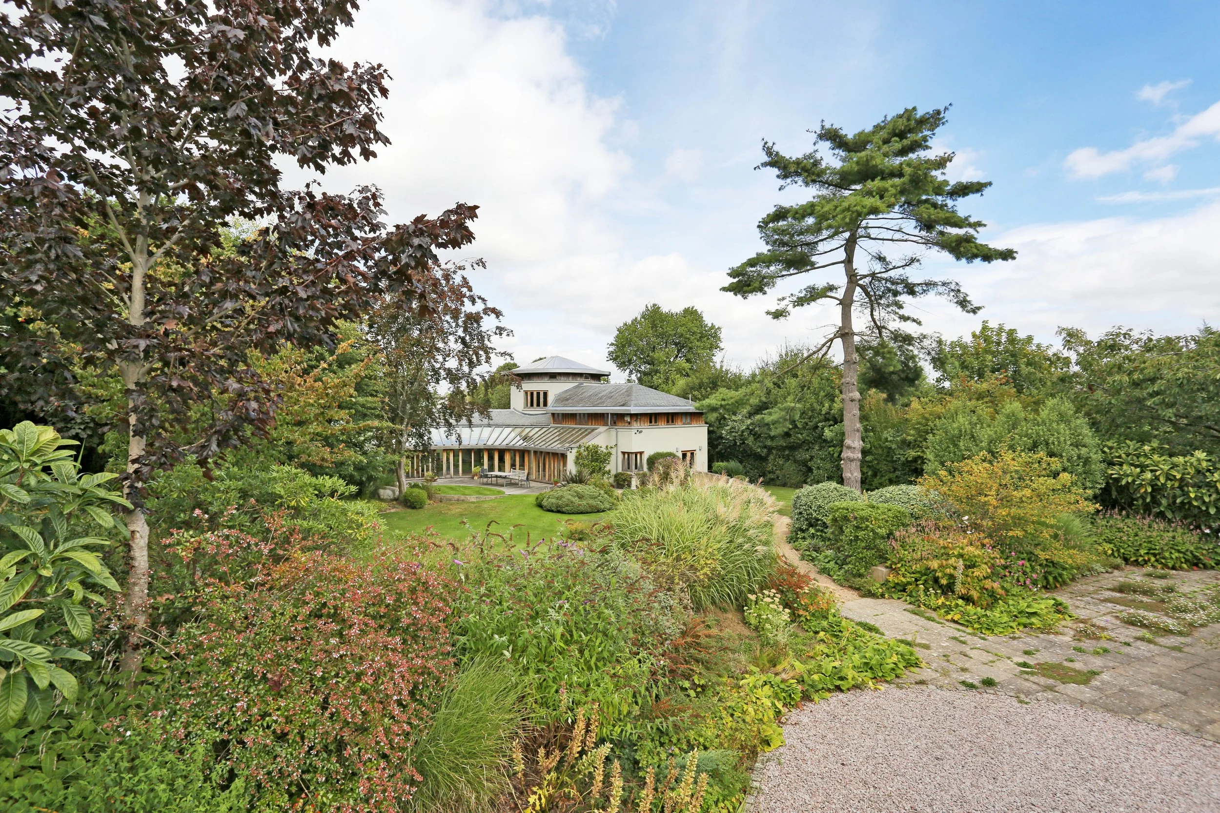 Lush botanical garden with flowering plants, manicured shrubs, mature trees, and a modern glass-roofed building beneath a blue sky