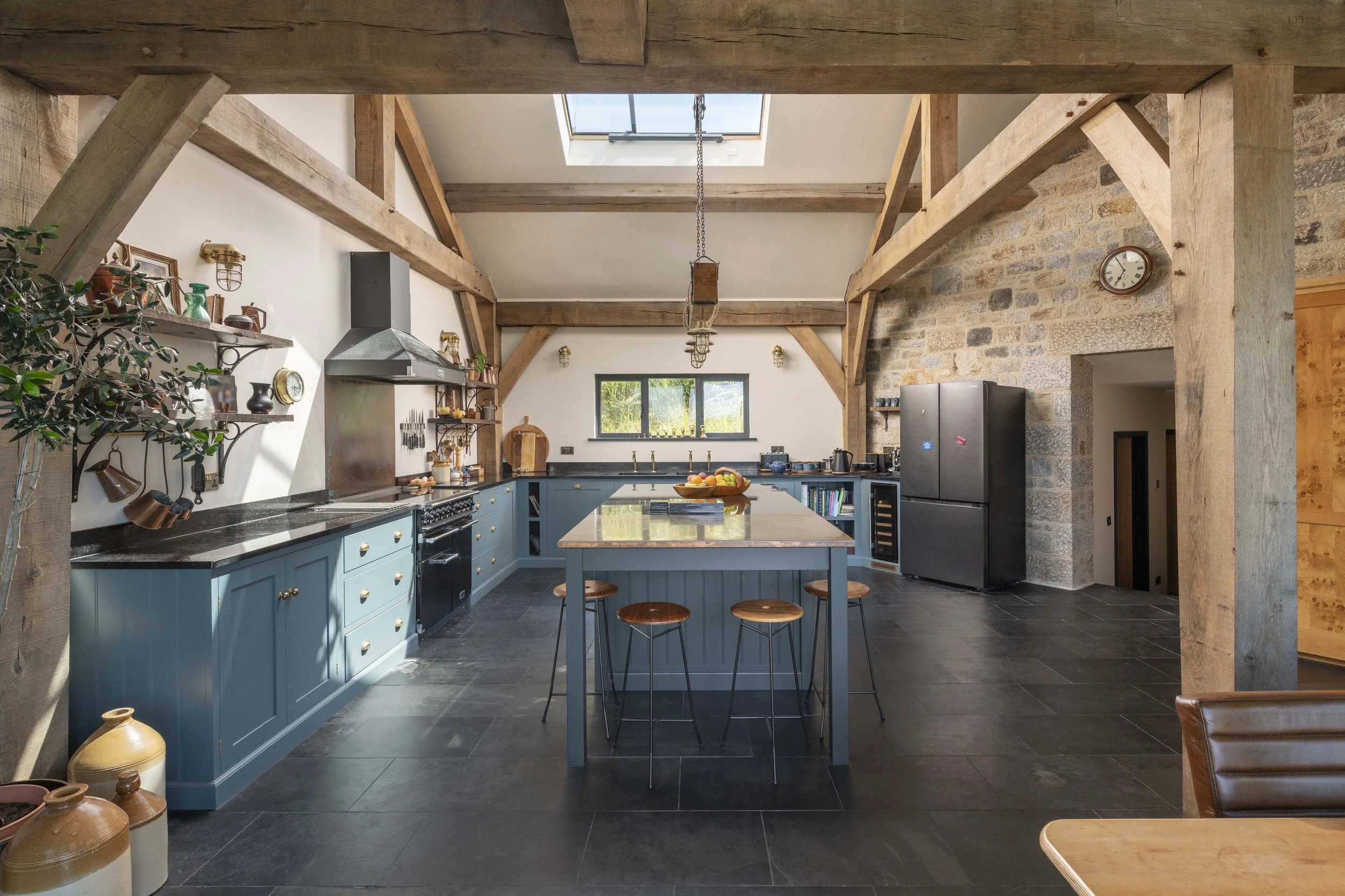 A spacious kitchen with a blue island counter, black countertops, wooden shelves with decorative items, a large window, and exposed wooden beams on the ceiling.