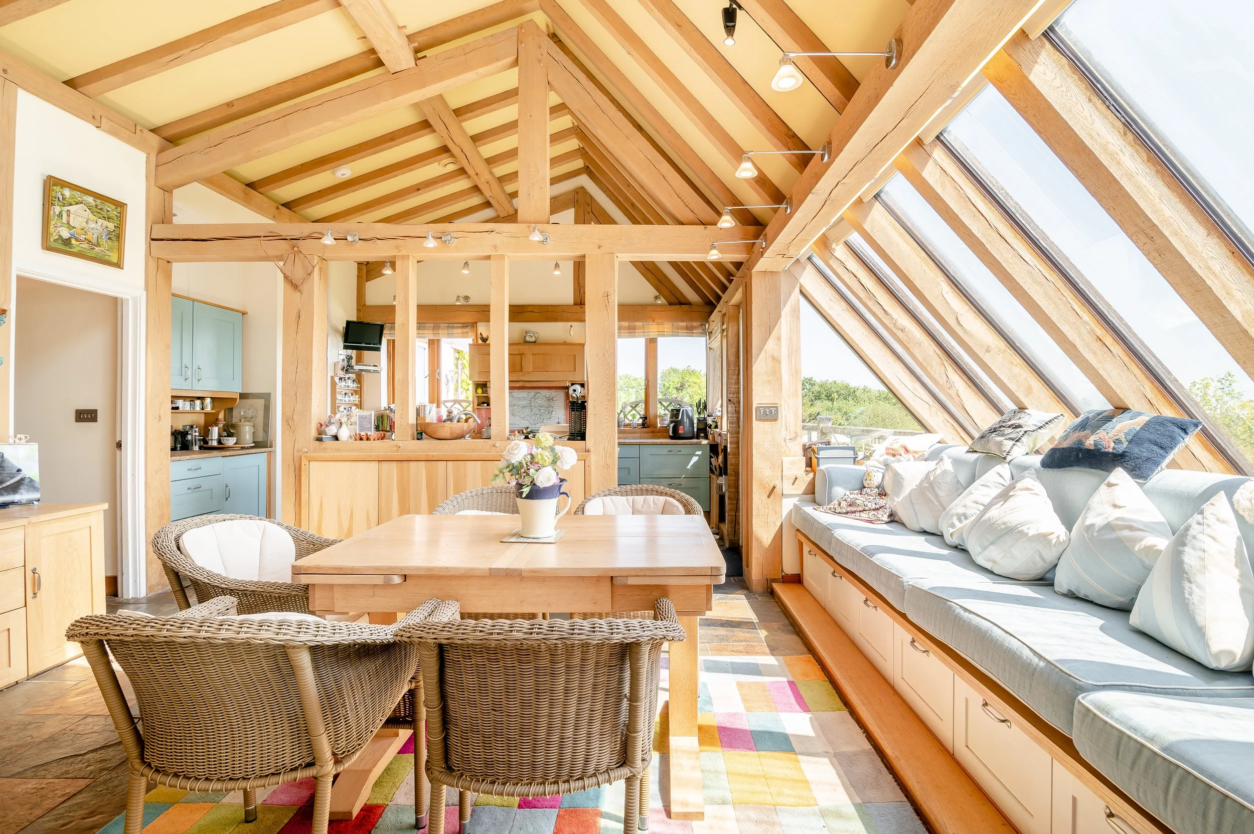 Bright living room with large angled windows, wooden beams, a sofa with pillows, a dining table with four wicker chairs, a colorful rug, and visible kitchen area with cabinets and appliances, all illuminated by natural sunlight.
