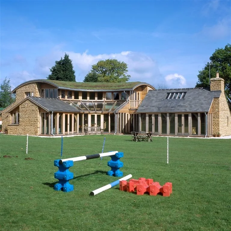 Large modern house with multiple sections, large windows, and a chimney on a grassy lawn. In the foreground, there are obstacle course pieces including a jump bar, hurdles, and red blocks. The sky is mostly clear with some clouds.