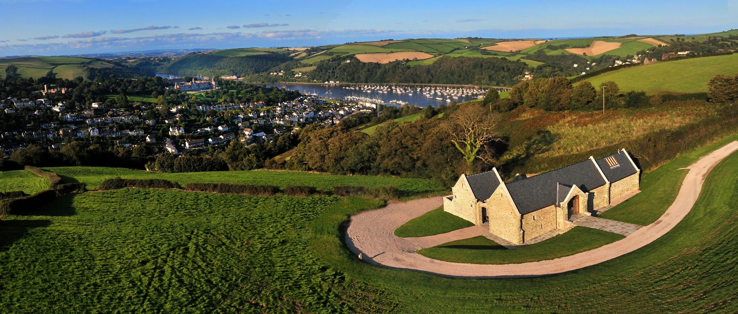 A scenic view of a countryside with a stone house, green hills, a town with houses, and a river with sailboats, under a partly cloudy sky.