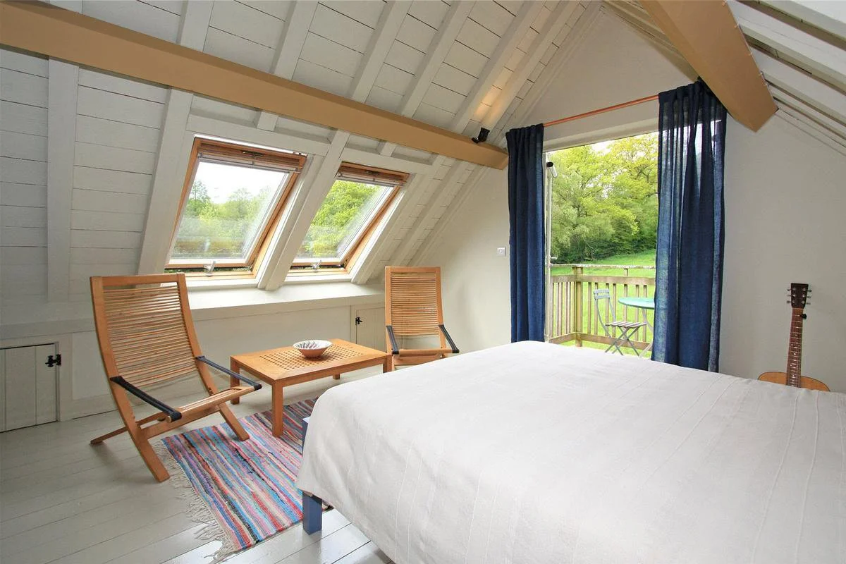 A cozy bedroom with a slanted ceiling and two skylight windows showing greenery outside. There's a bed with a white cover, a guitar leaning against the wall, two wooden chairs, a small wooden table with a bowl, a colorful striped rug, and a sliding g